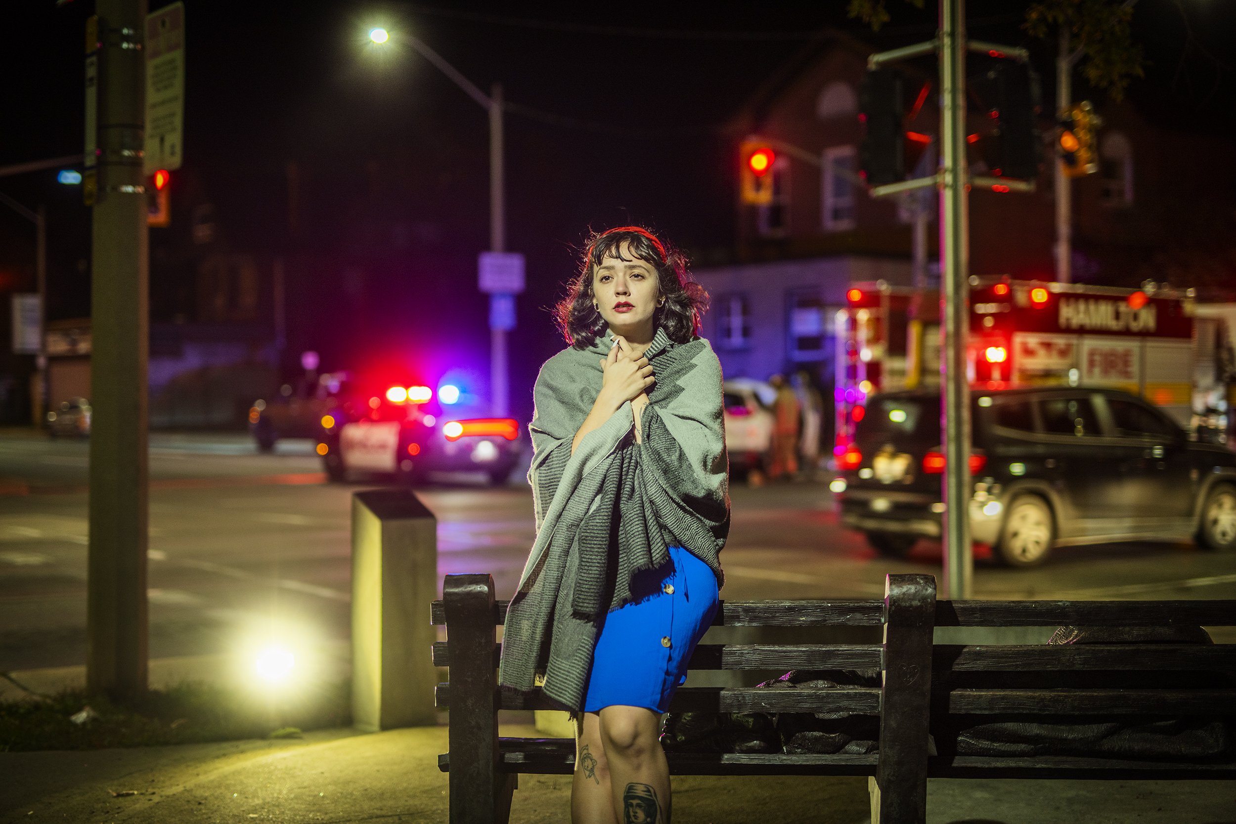 A woman with dark hair, tattoos on her leg, and wearing a blue dress and striped shawl, standing on a night street near police car and fire truck with flashing lights.