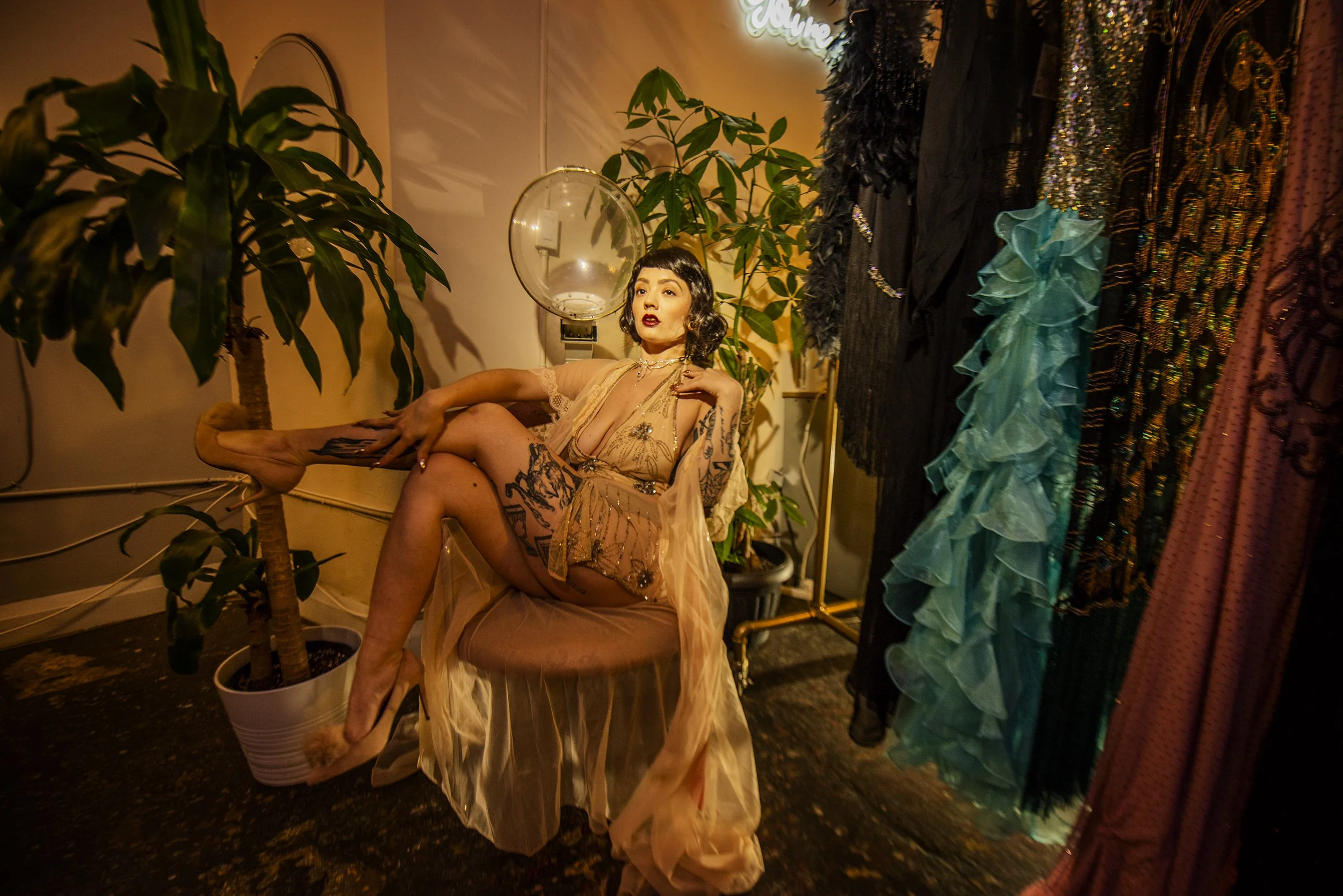 A woman with tattoos, dressed in a sheer peach-colored dress, sitting in a room decorated with plants and clothing racks. She has dark hair styled in vintage waves and red lipstick. The room has warm lighting and a neon sign that reads ‘Home’ on the 
