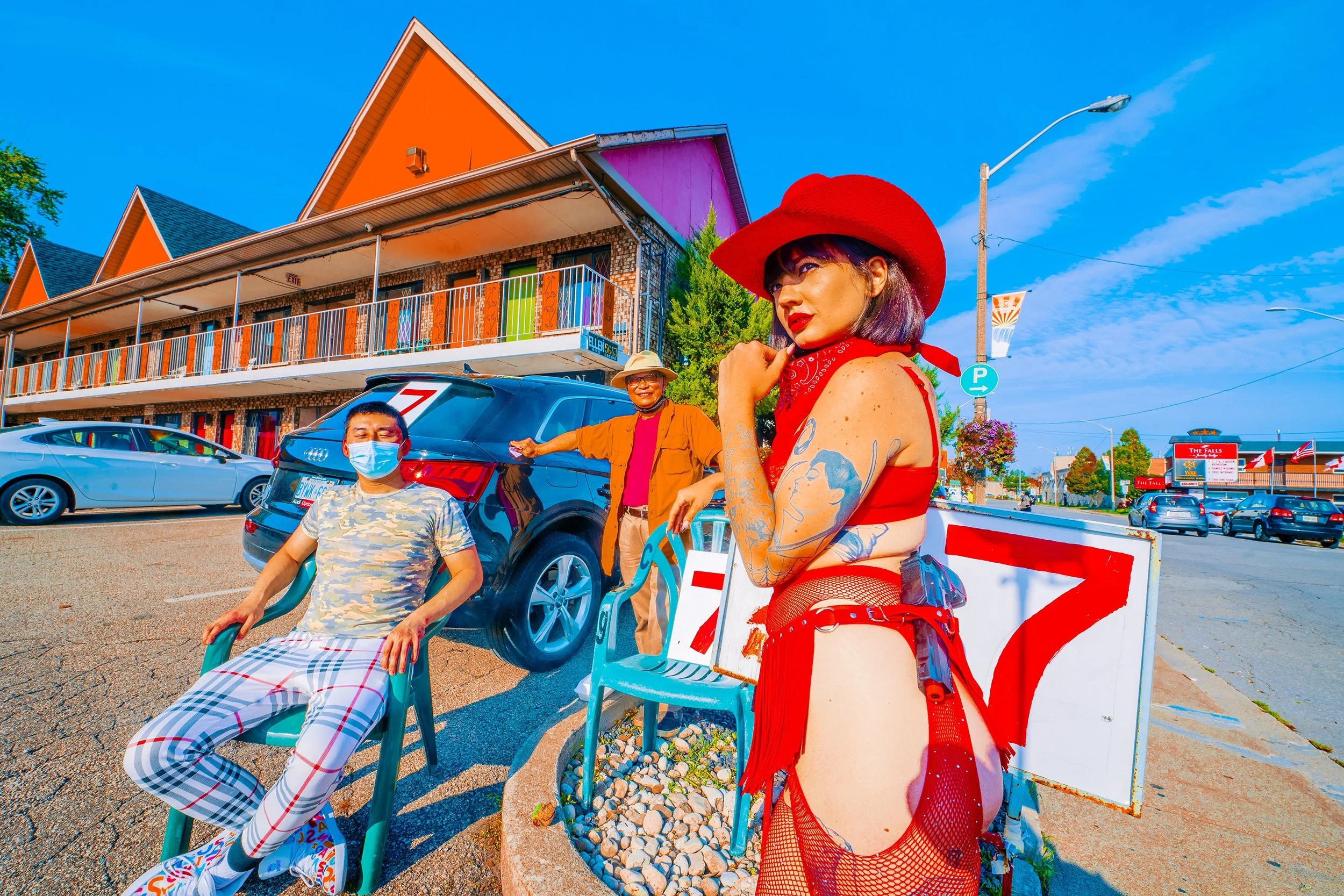 People standing and sitting outside near parked cars on a sunny day, with colorful buildings and signs in the background.