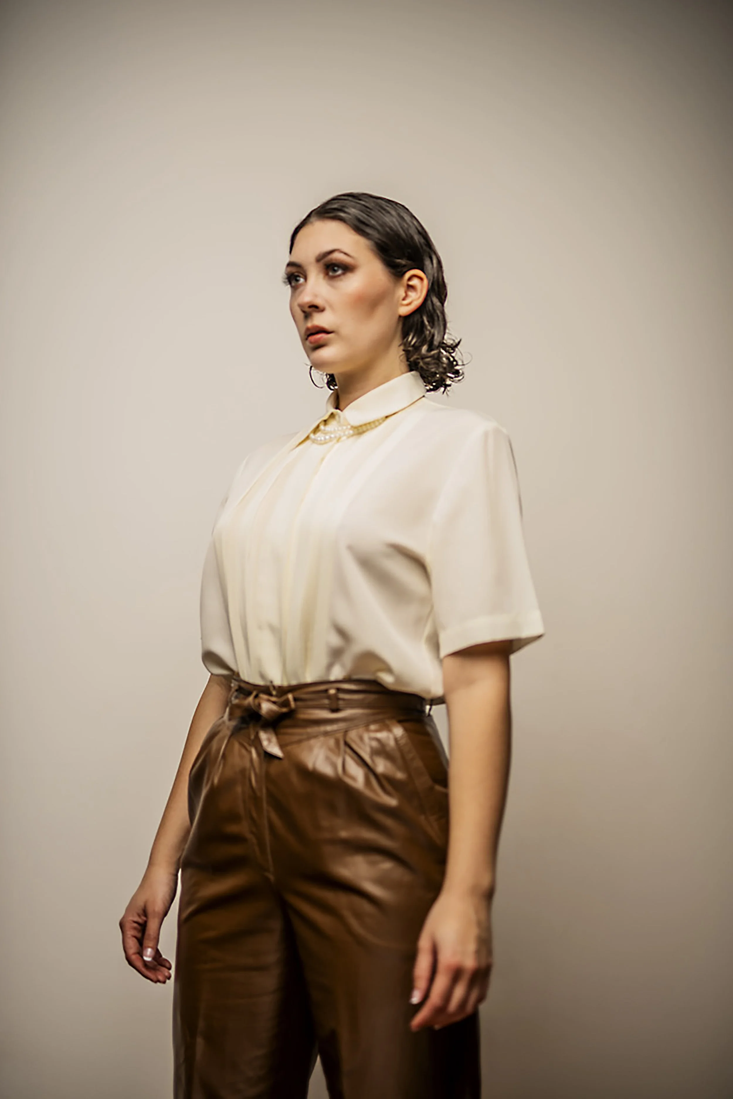 A woman with short, dark wavy hair wearing a cream-colored blouse with a collar and a pearl necklace, paired with brown high-waisted pants, standing against a plain background.