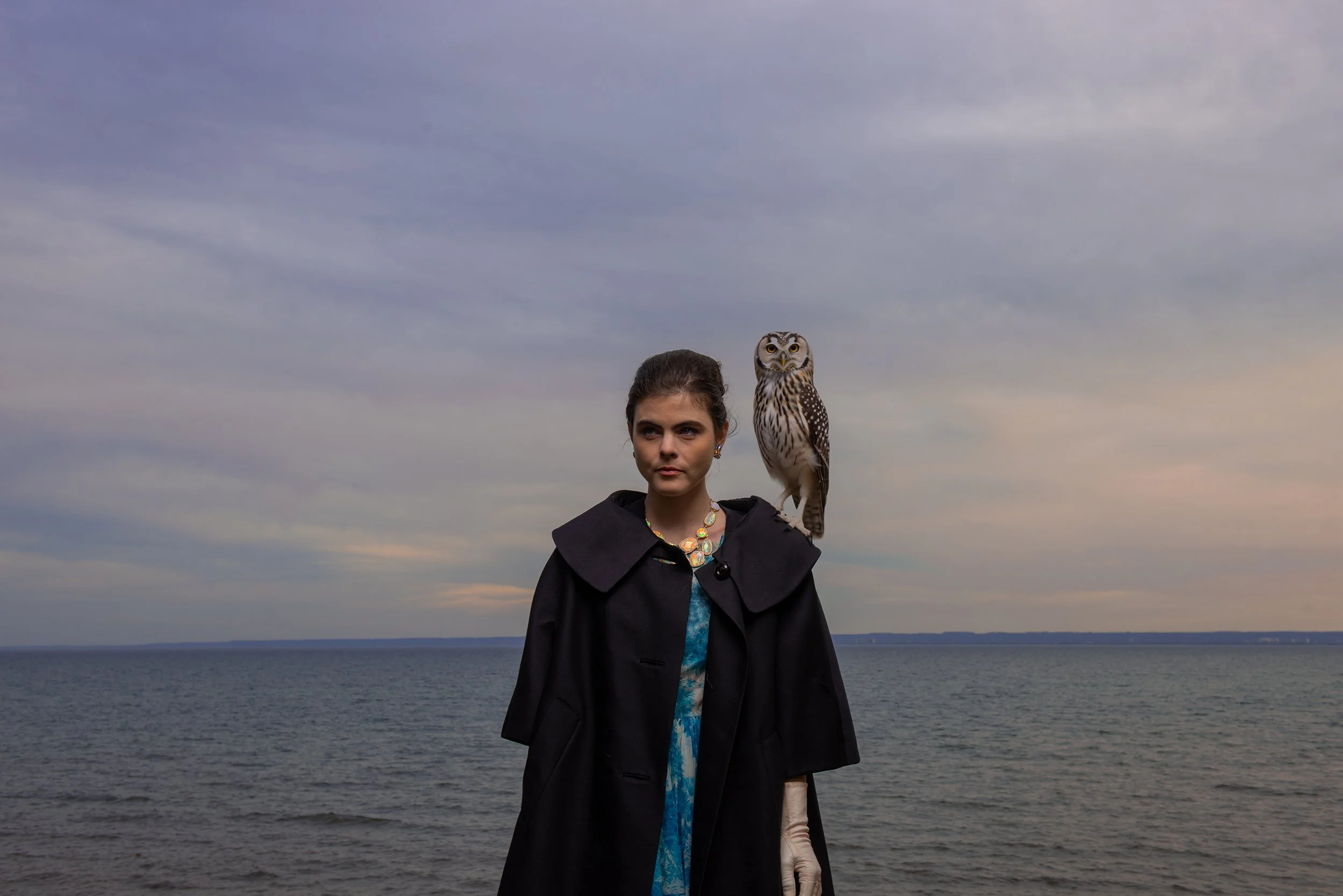 A woman standing by a body of water with a snowy owl perched on her shoulder, against a cloudy sky at dusk.