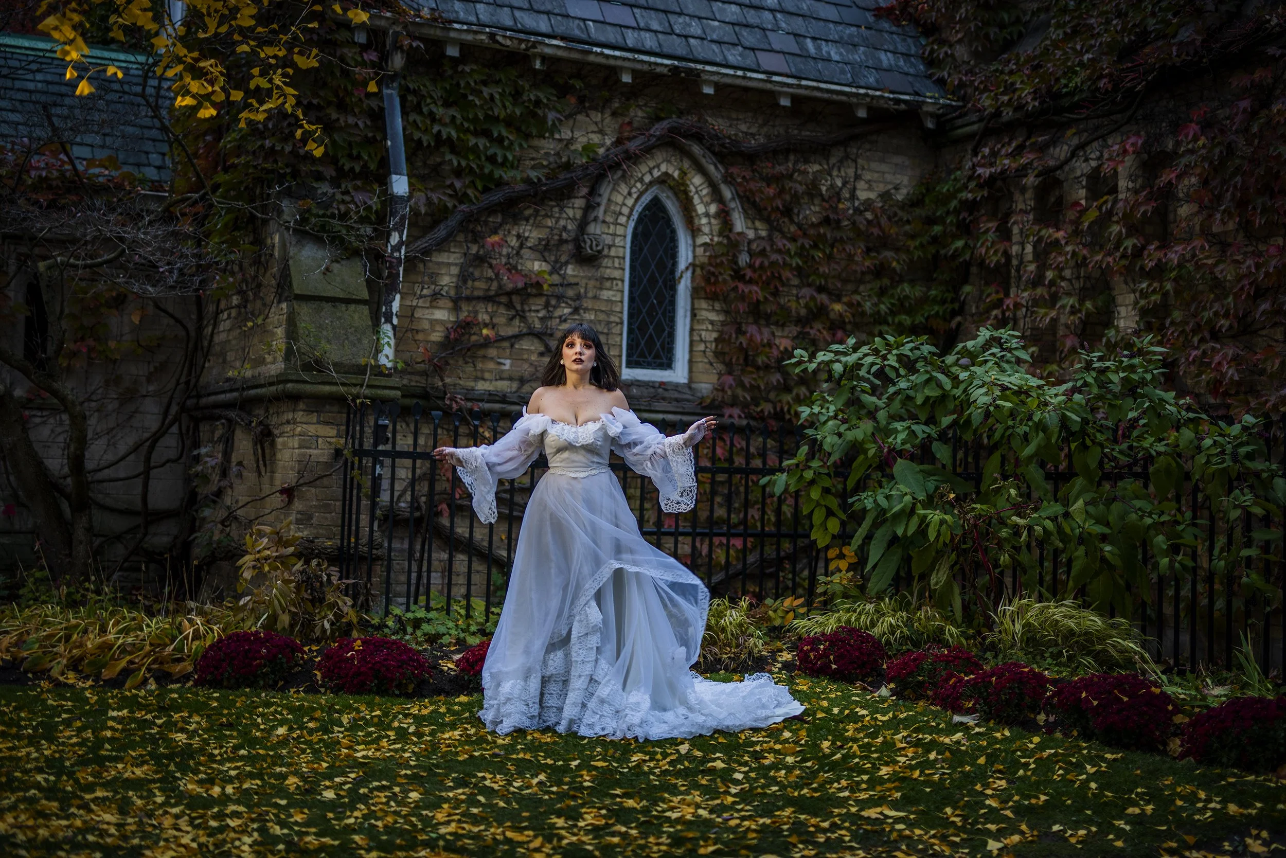 A woman dressed in a white vintage wedding gown with lace sleeves, standing in a garden with yellow and red fallen leaves, in front of a brick building covered in climbing ivy.