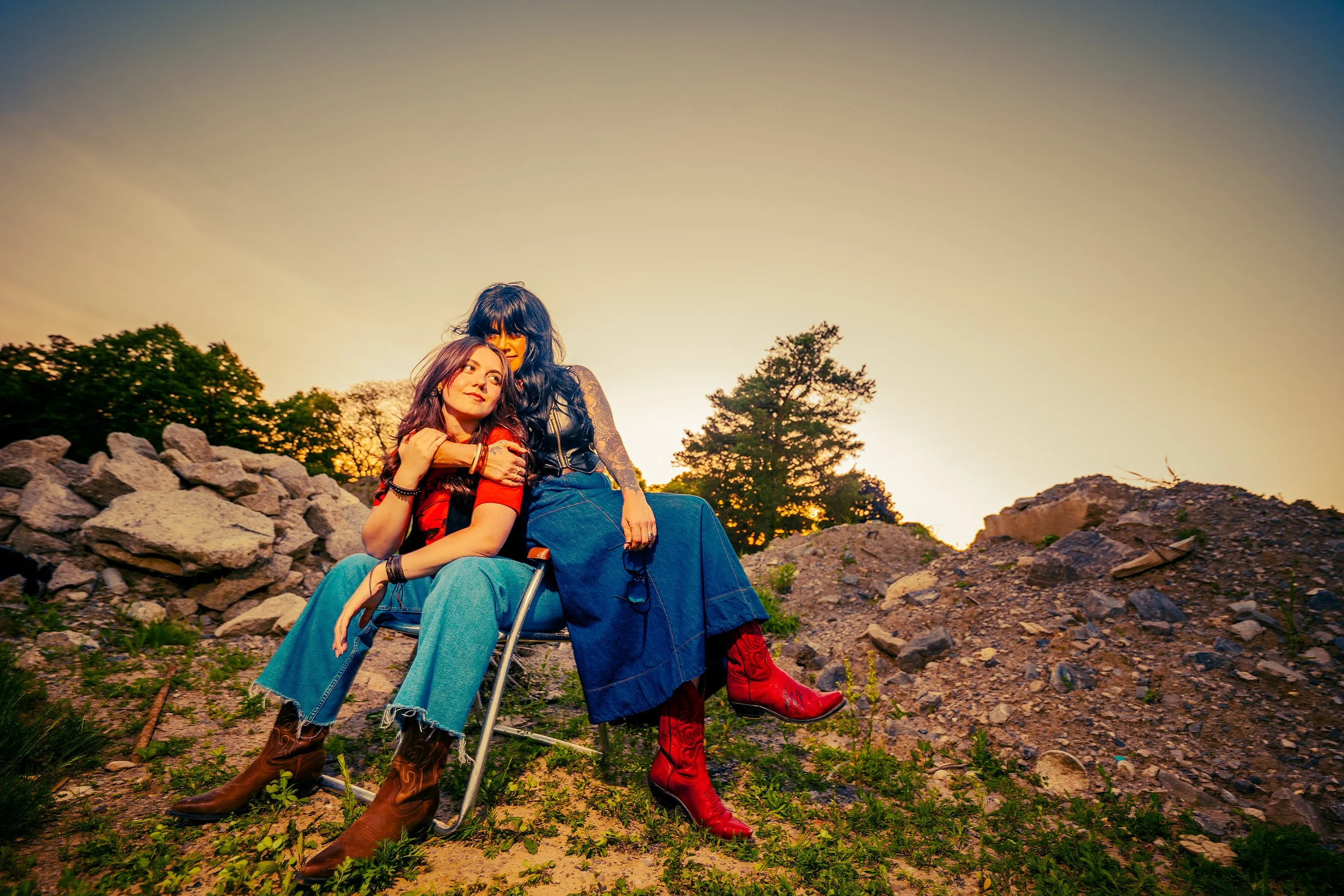Two women sitting outdoors on rocks during sunset, embracing each other, one in a wheelchair, surrounded by trees and rocks.