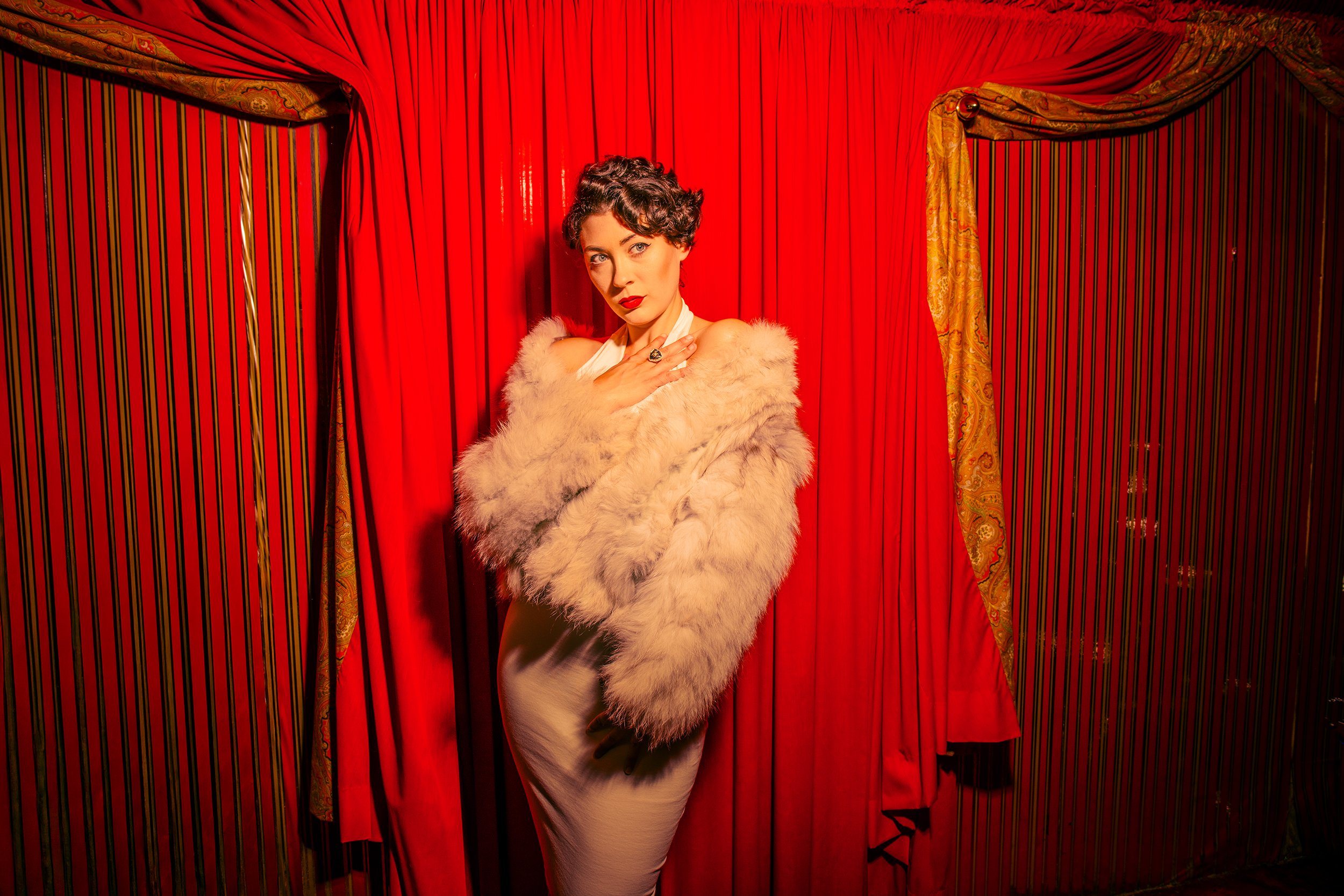 A woman with short curly dark hair and red lipstick posing in front of red curtains, wearing a faux fur coat and a white dress with a black belt.