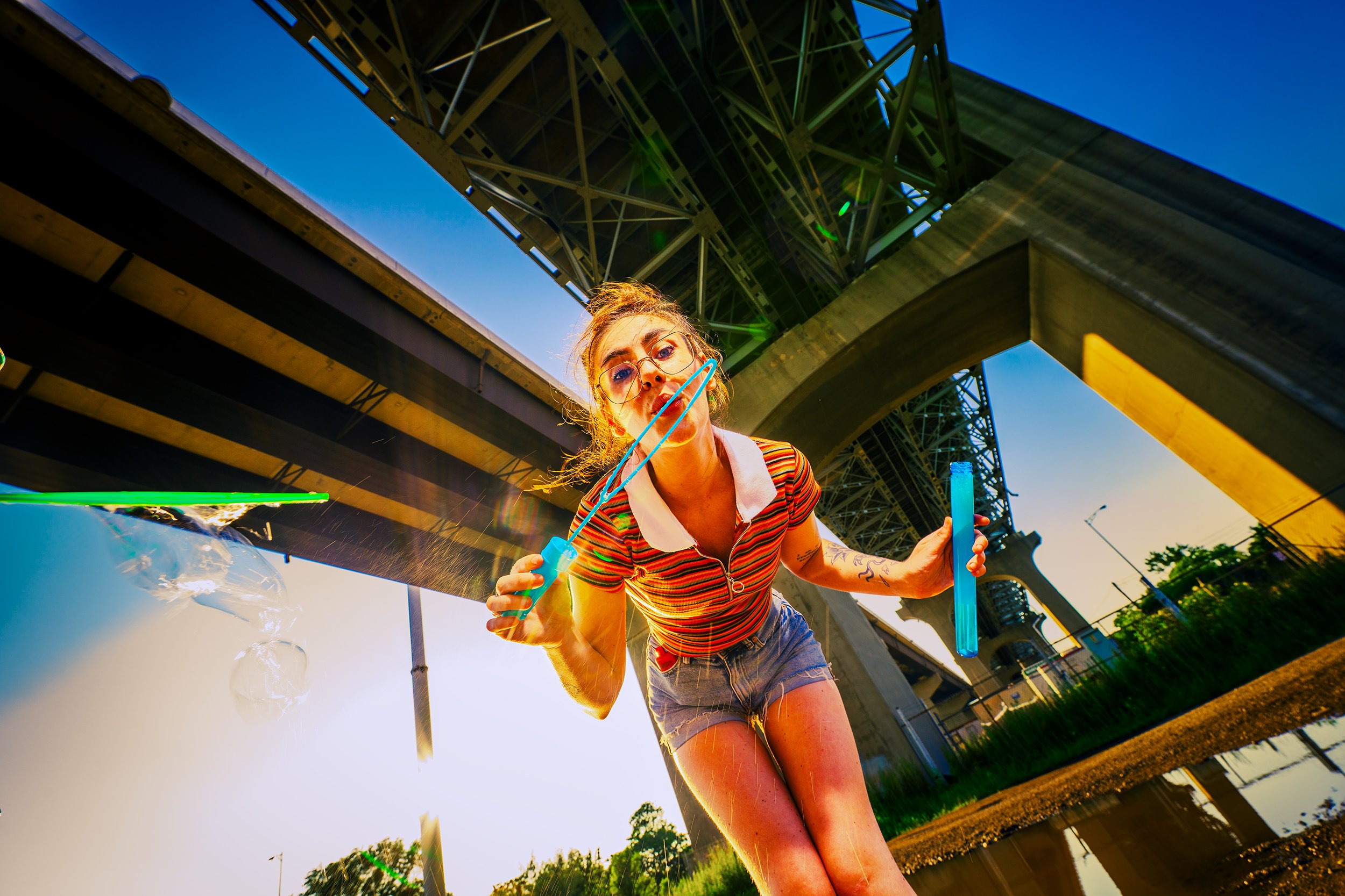 A woman in a striped shirt and glasses blowing bubbles outdoors under a bridge during sunset.