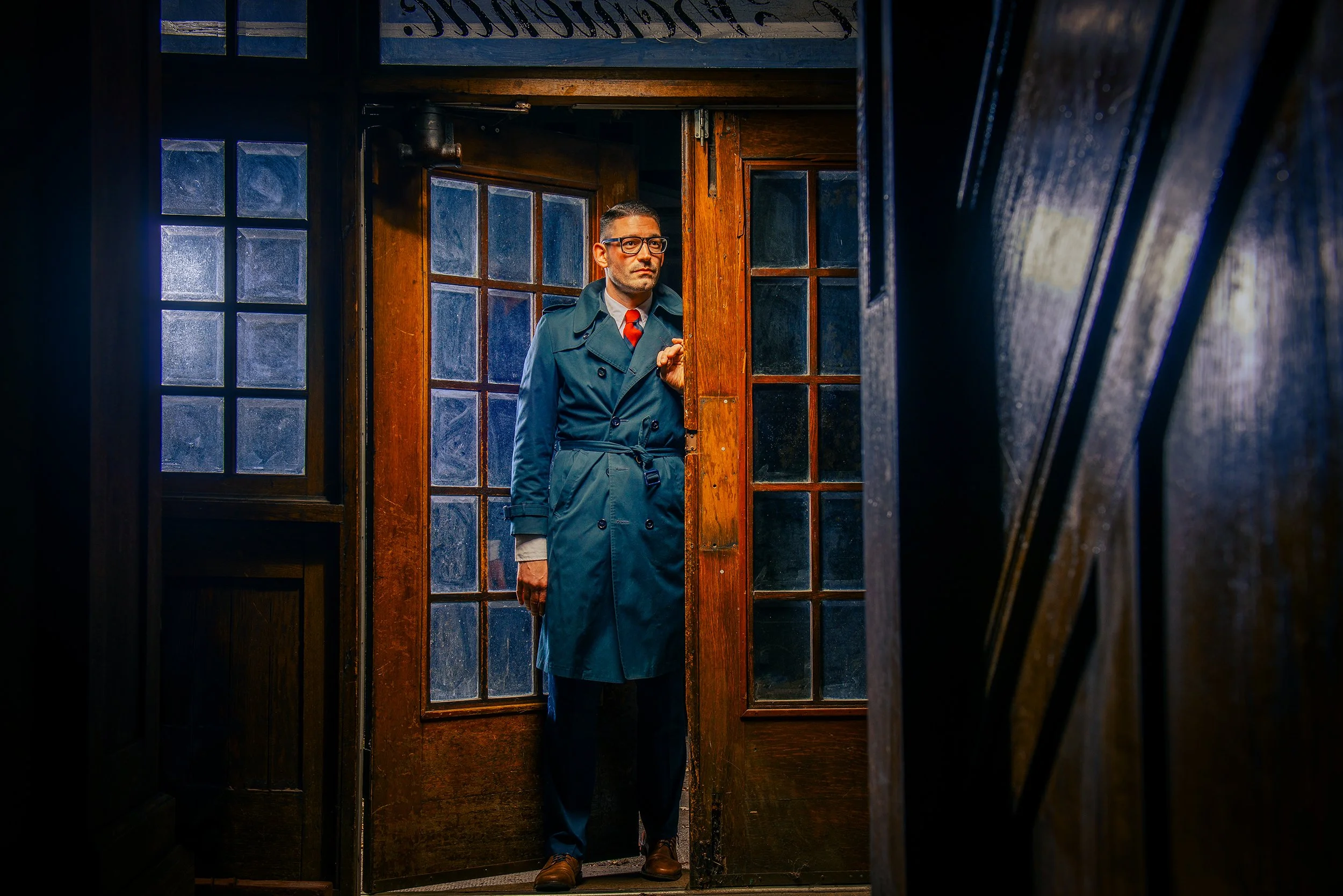 A man wearing glasses, a blue trench coat, and a red tie standing partially behind a wooden doorframe, looking out with a serious expression in a dimly lit interior with wooden and glass elements.