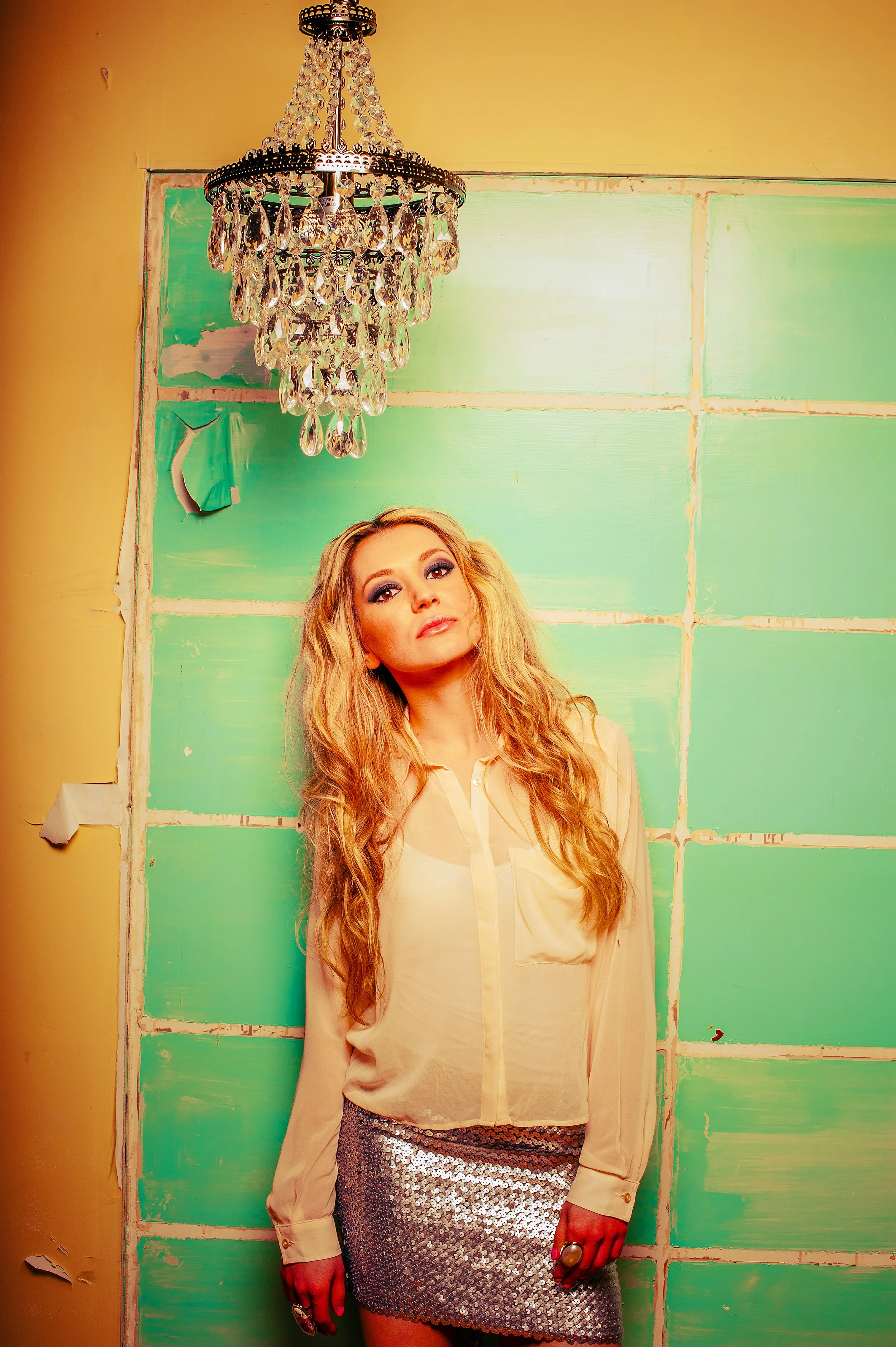 Woman with long blonde hair standing in front of a green painted wall and a chandelier, wearing a beige blouse and a sparkly silver skirt.