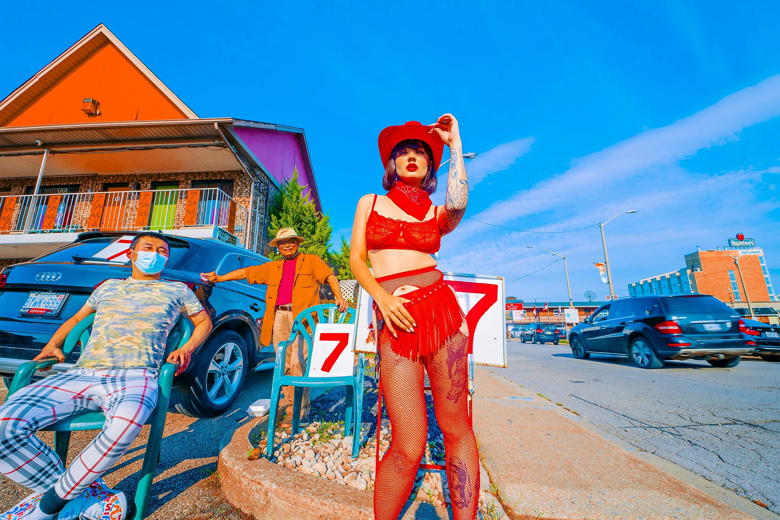 Street scene with three people, one woman dressed in red lingerie and fishnet stockings wearing a large red hat, standing near a sign with a red '7' on it, while two casually dressed men sit nearby on a sunny sidewalk; background features a building 