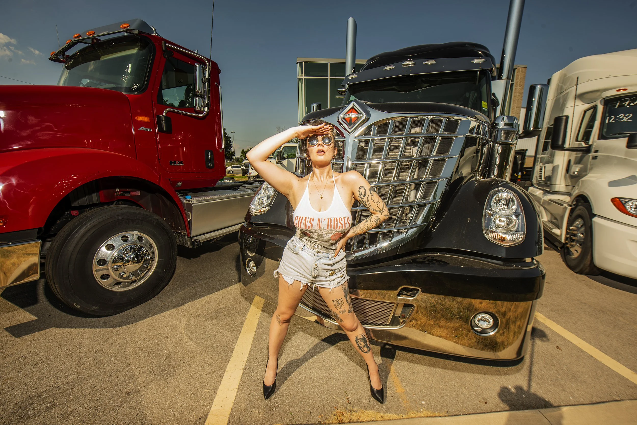 A woman with tattoos and wearing a Guns N' Roses tank top, denim shorts, black high heels, and sunglasses, standing with one hand on her forehead and the other on her hip, posing in front of a black semi-truck with a large chrome grille, parked in a 