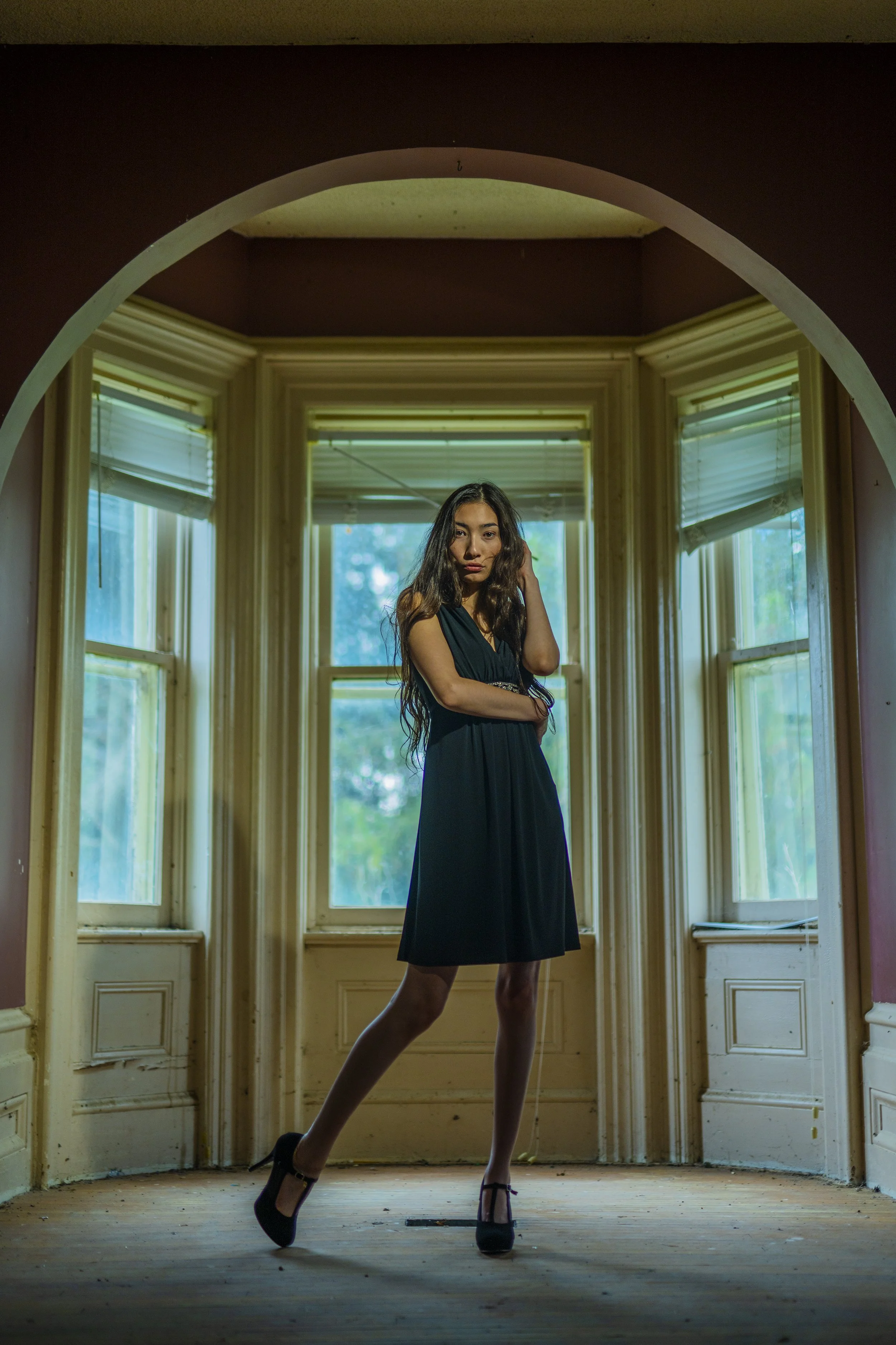 A woman in a black dress and high heels standing in front of windows in a dimly lit room.