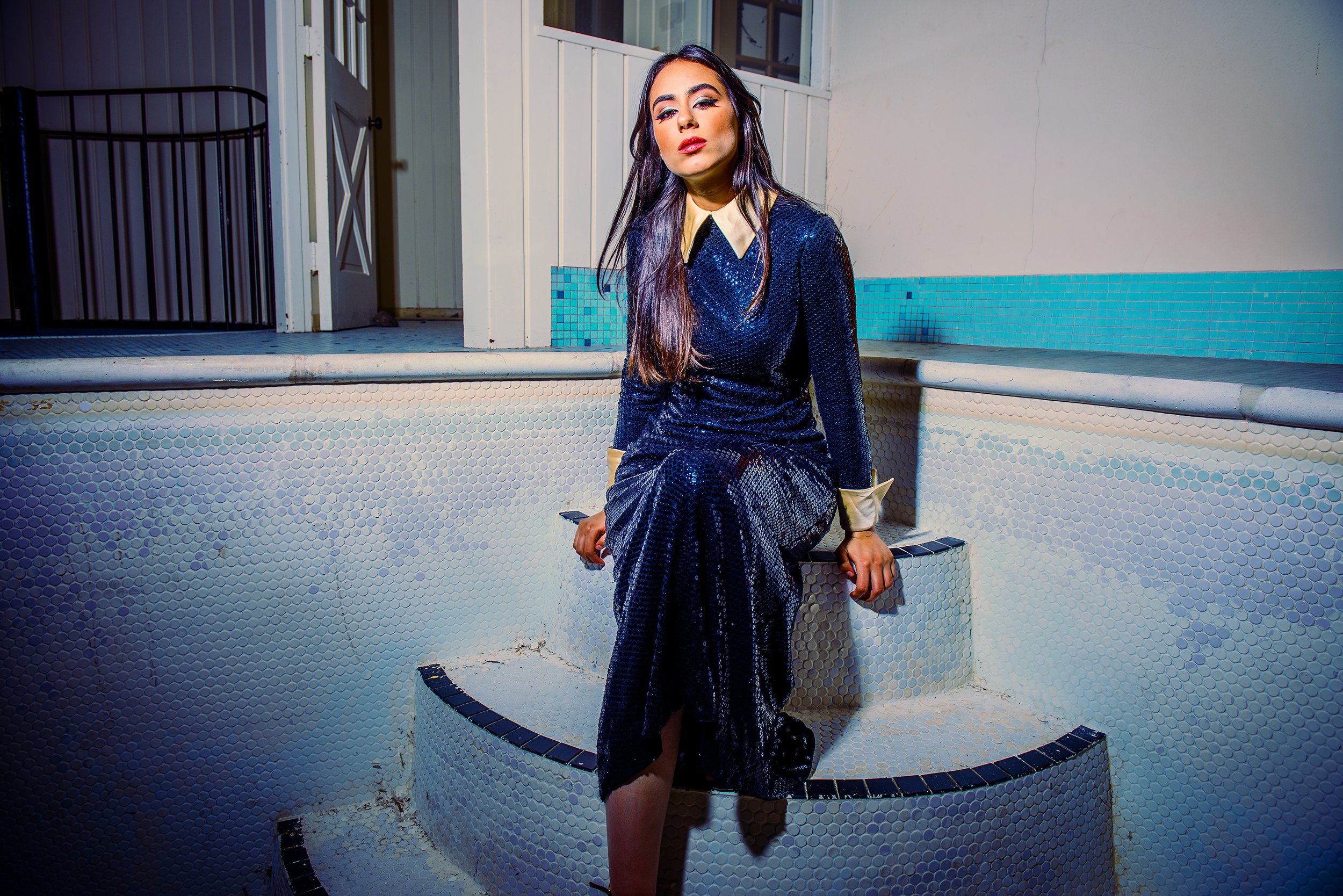 A woman with long dark hair and makeup sitting on the edge of an empty, tiled hot tub in an indoor setting.