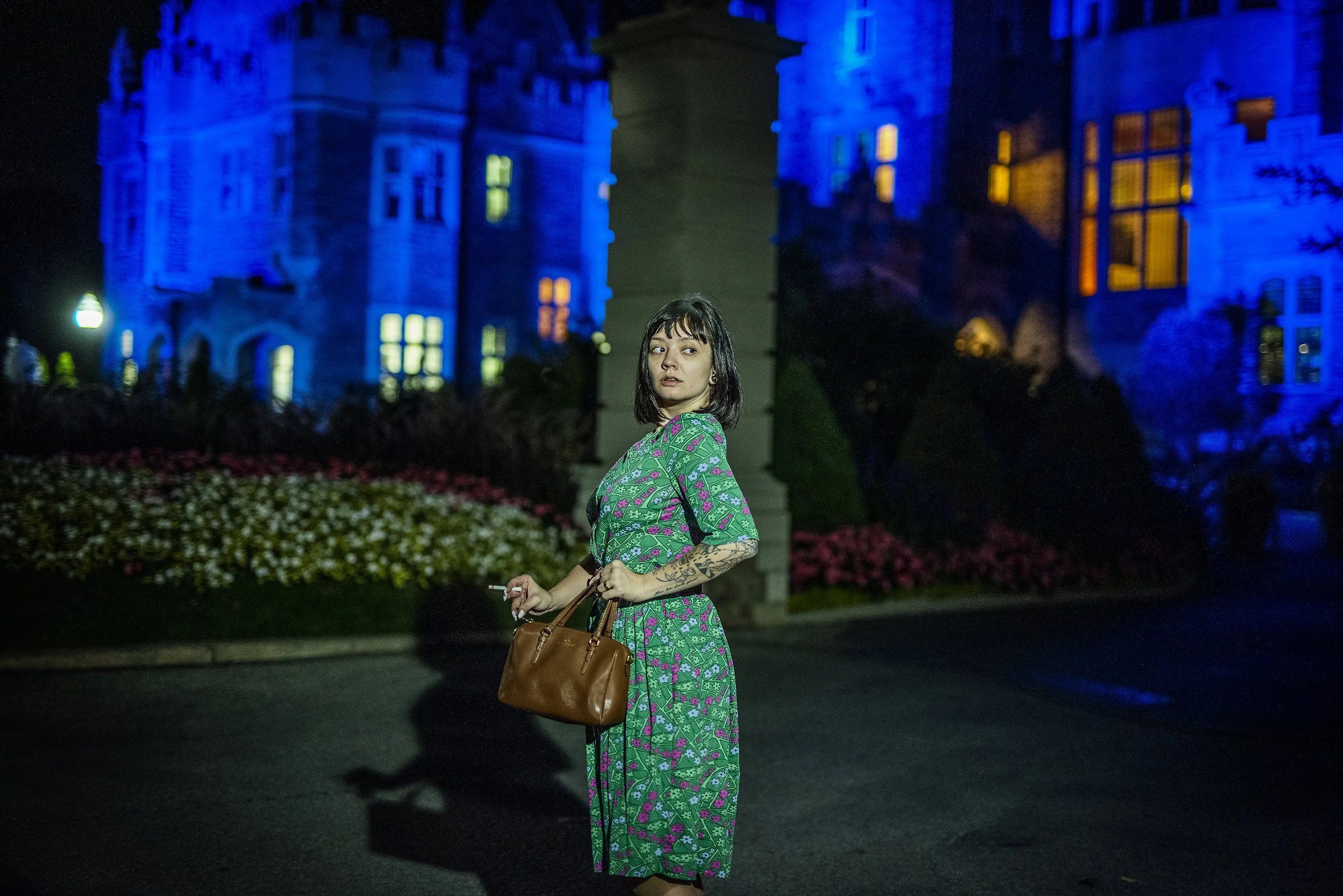 A young woman with dark hair and tattoos on her arm standing outside at night in front of a large, illuminated castle-like building with blue and yellow lights. She is wearing a long, green floral dress and carrying a brown handbag, holding a cigaret
