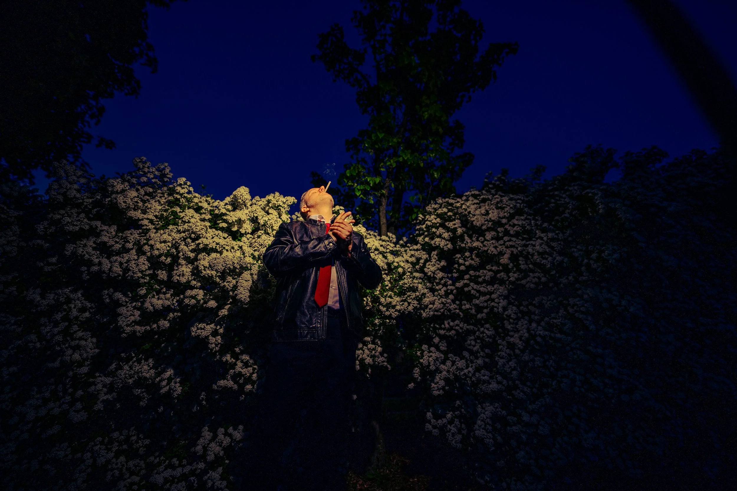 A man wearing a leather jacket and red tie standing outside at night, surrounded by flowering bushes, looking up with his hands clasped, smoking a cigarette.