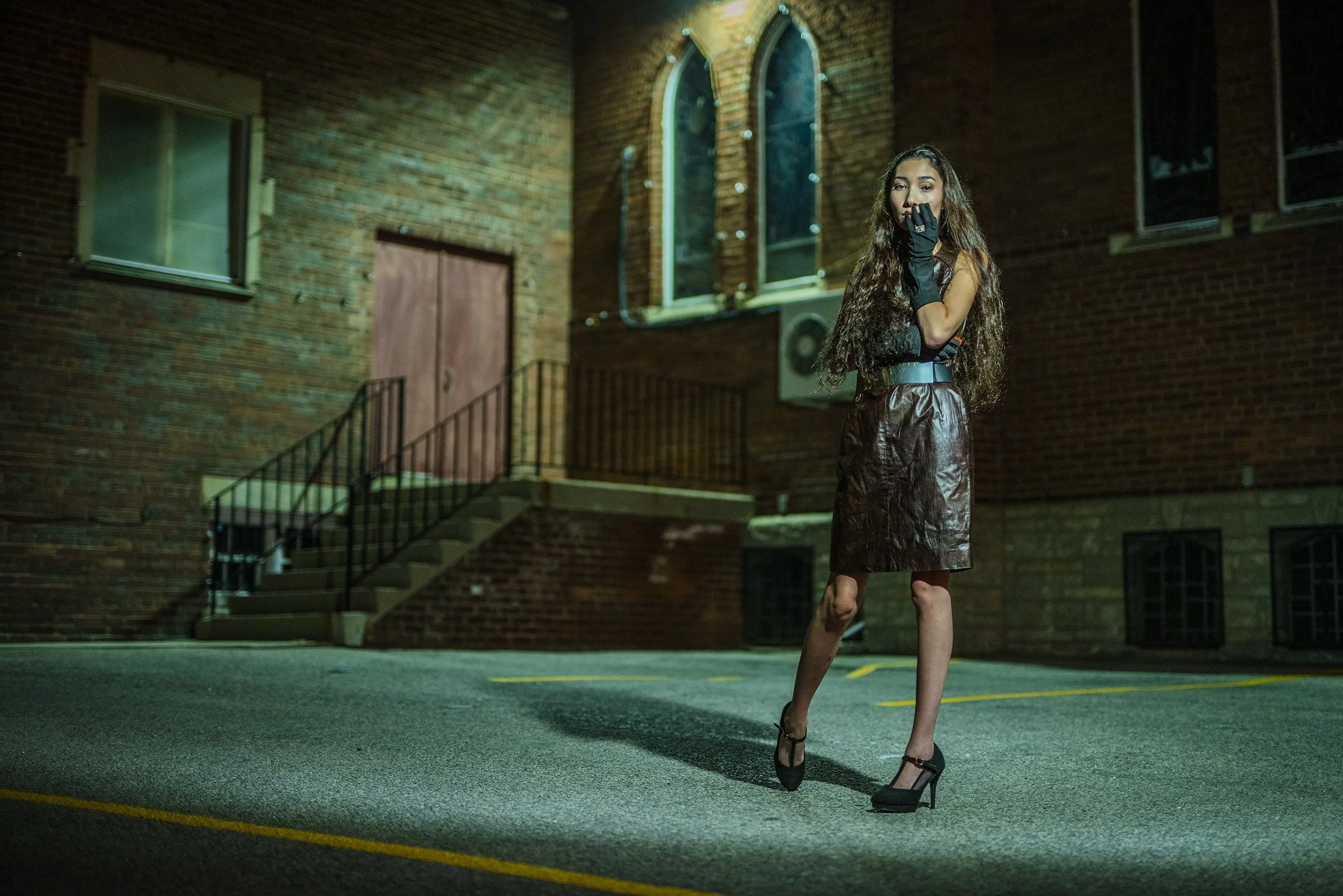 A woman standing outdoors at night in front of a brick building with arched windows. She has long wavy hair, is wearing a sleeveless top, a shiny brown skirt, and black high heels. She covers her mouth with her right hand, which is gloved in black.