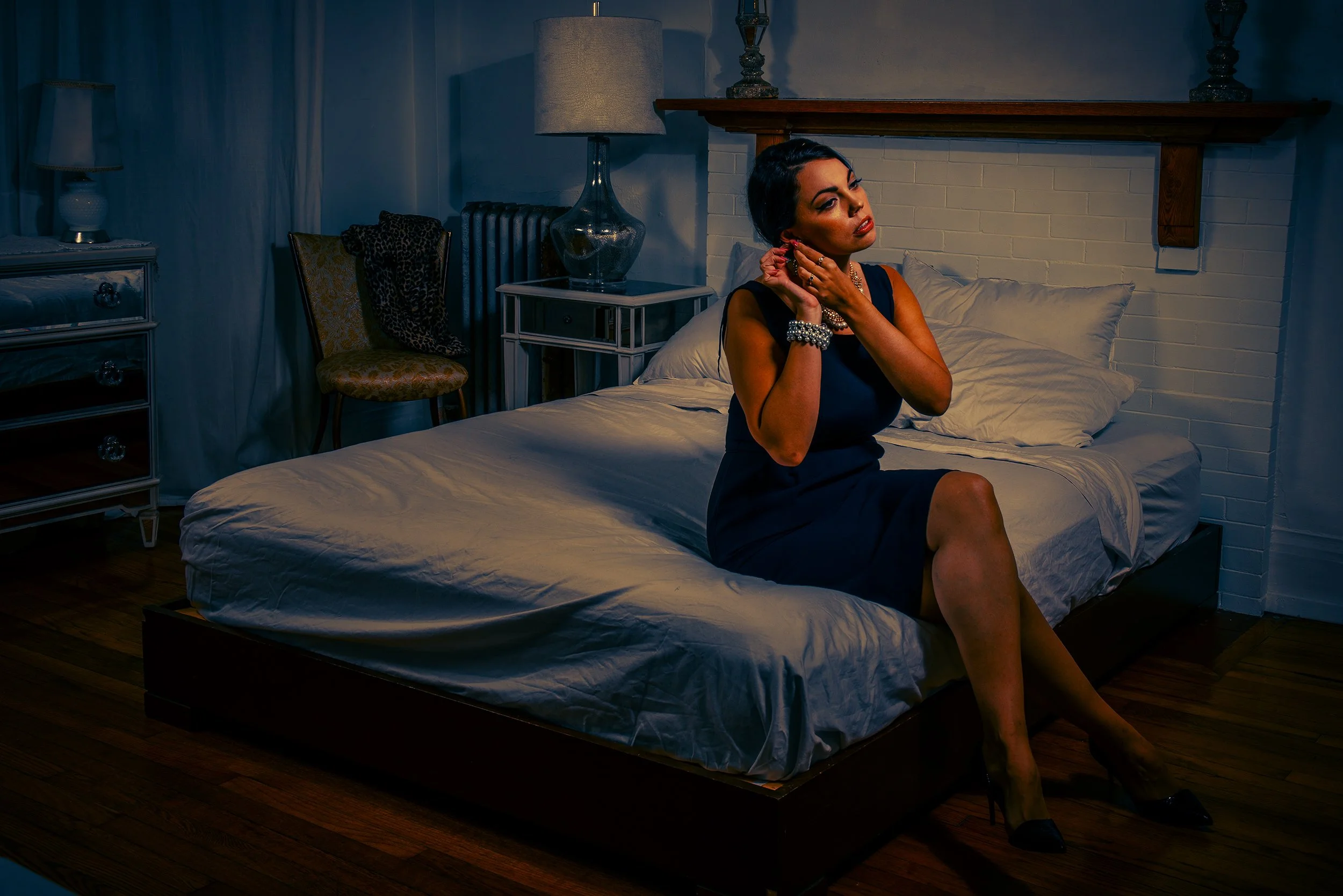 A woman with dark hair and makeup wearing jewelry sits on a bed in a dimly lit bedroom, touching her earrings and looking to the side.