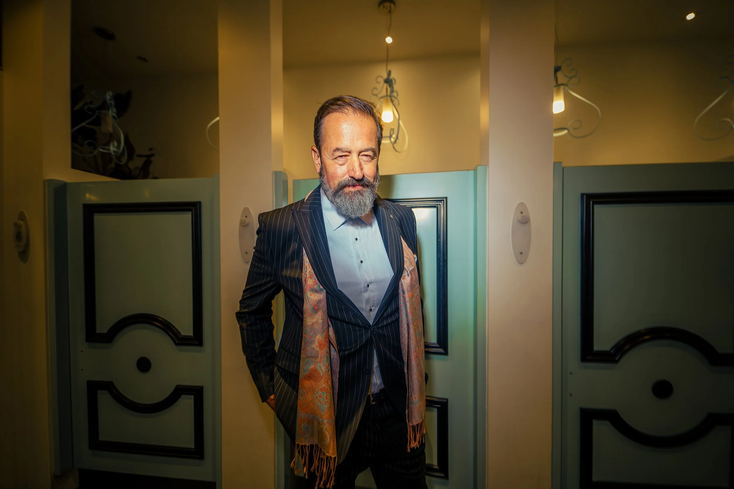 A man with a beard and mustache wearing a dark pinstripe suit and a colorful scarf, standing in an art gallery or exhibition space with yellow walls and framed artwork.