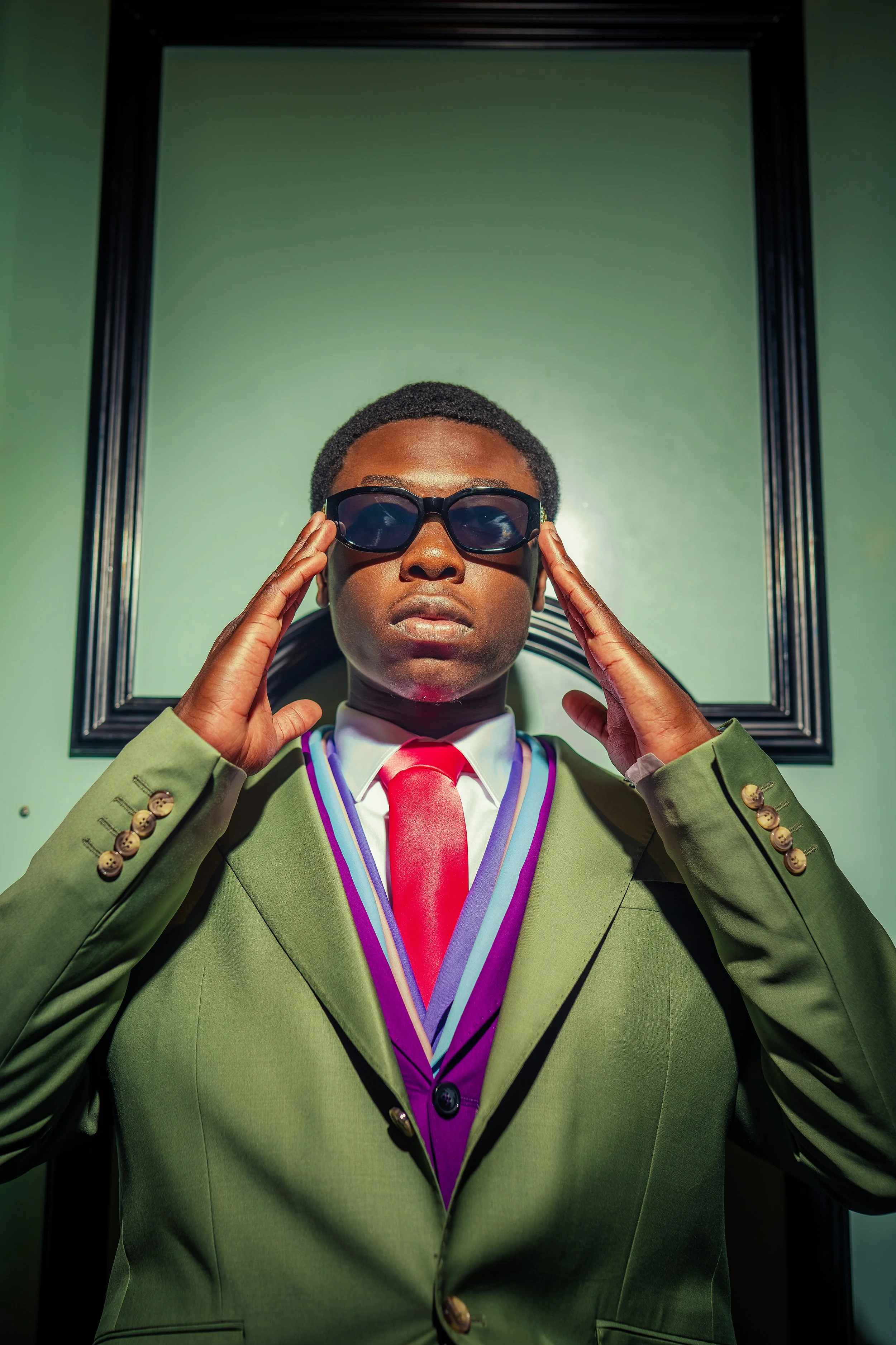 A man wearing a green suit, red tie, and jewelry, adjusting black sunglasses, standing in front of a green wall with black framing.