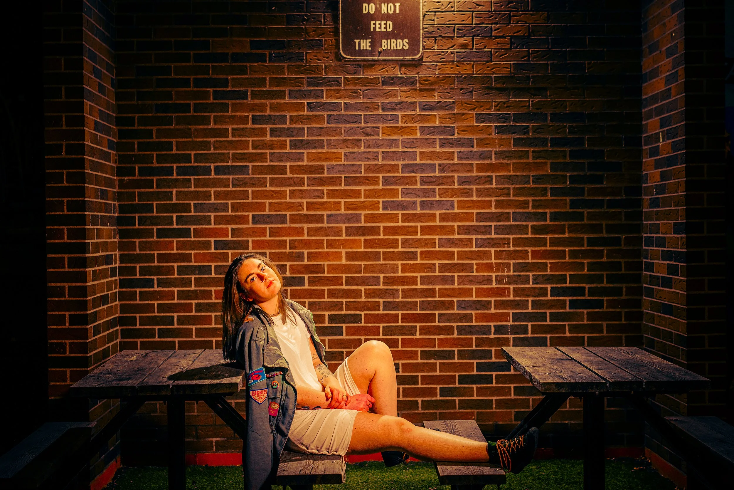 A young woman with long brown hair, tattoos, and wearing a gray jacket and boots, is sitting on a wooden picnic table with her legs extended and crossed at the ankle. She is wearing a white t-shirt and shorts. Behind her, a brick wall features a sign
