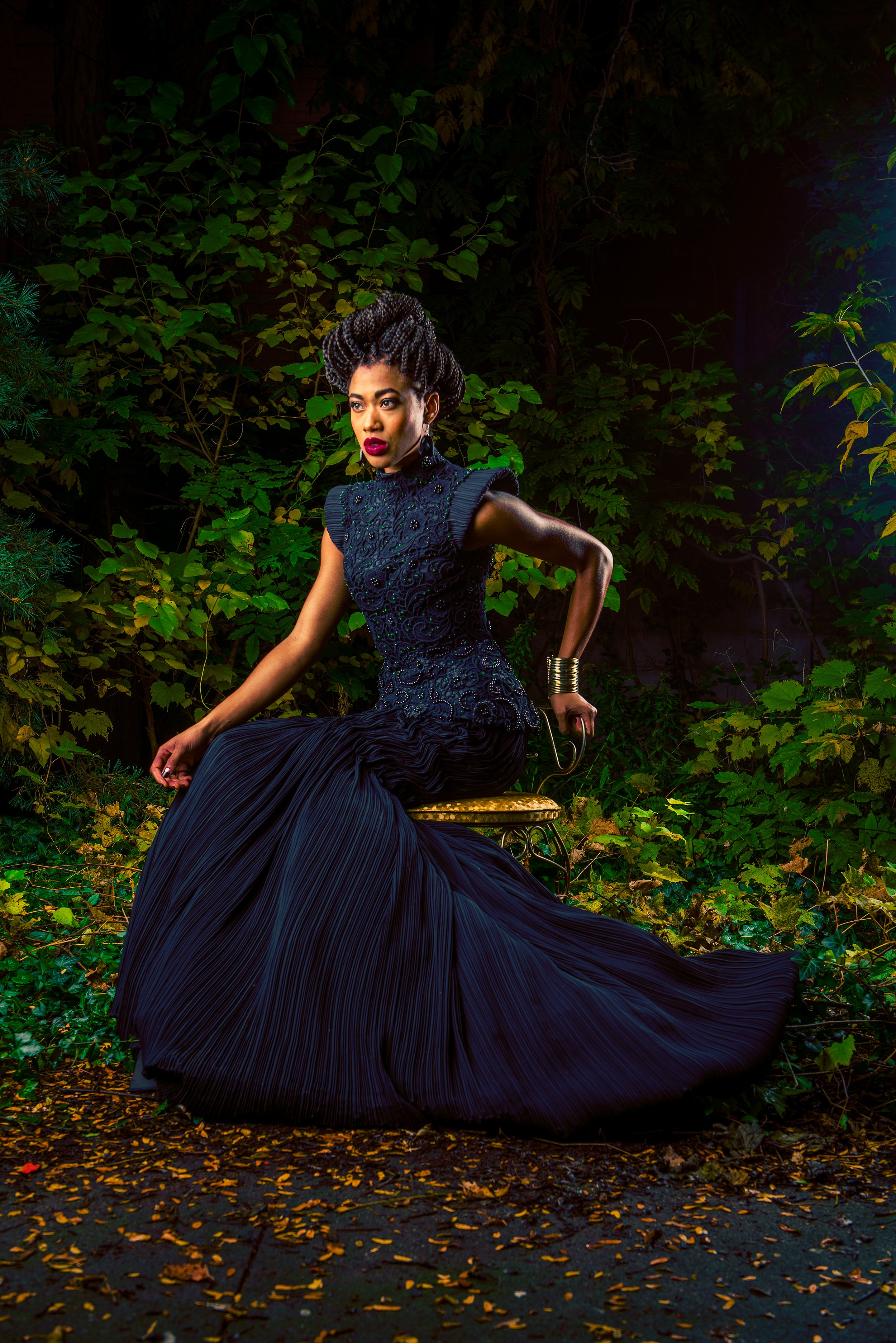 A woman with styled hair wearing a detailed black dress sitting outdoors on a gold-colored stool with fallen leaves on the ground and green leafy plants behind her.