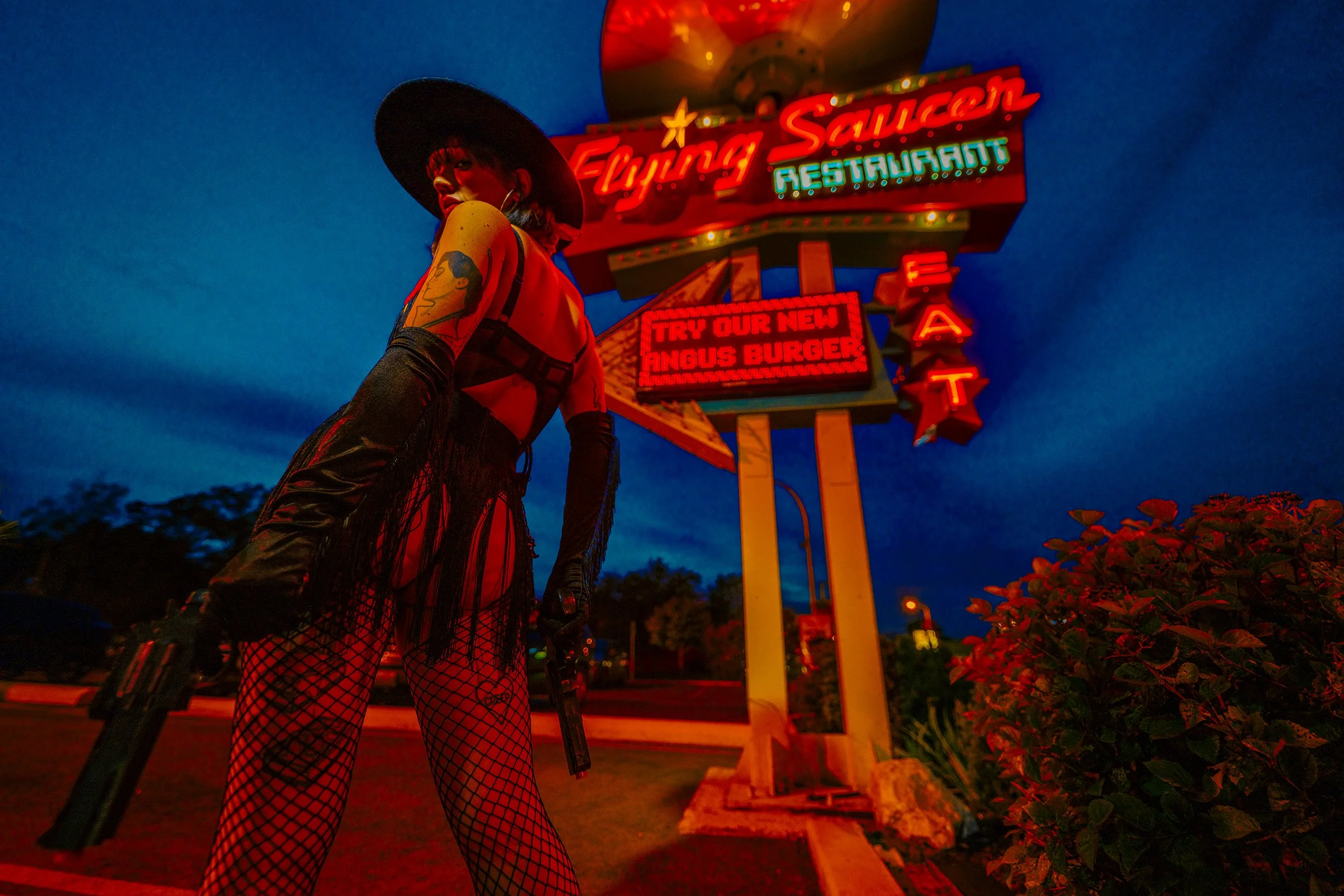 A person dressed in a black fringed outfit with fishnet stockings, black gloves, and a wide-brimmed hat, holding a handgun, standing in front of a brightly lit neon sign at night. The sign reads 'Flying Saucers Restaurant' and advertises a new Angus 