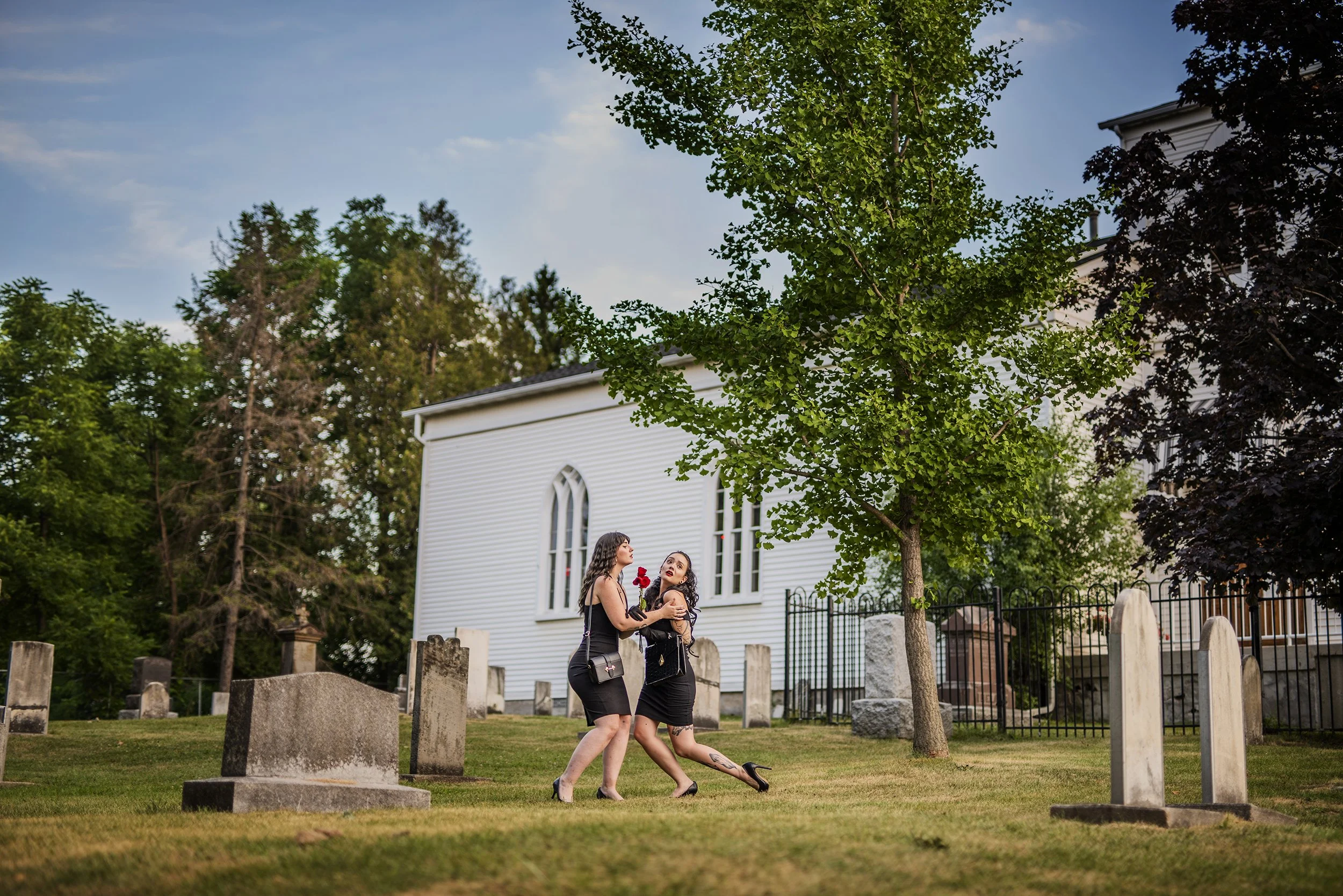 Two women in black dresses and high heels dancing and laughing in a cemetery with gravestones, a white church in the background, and trees around during the daytime.