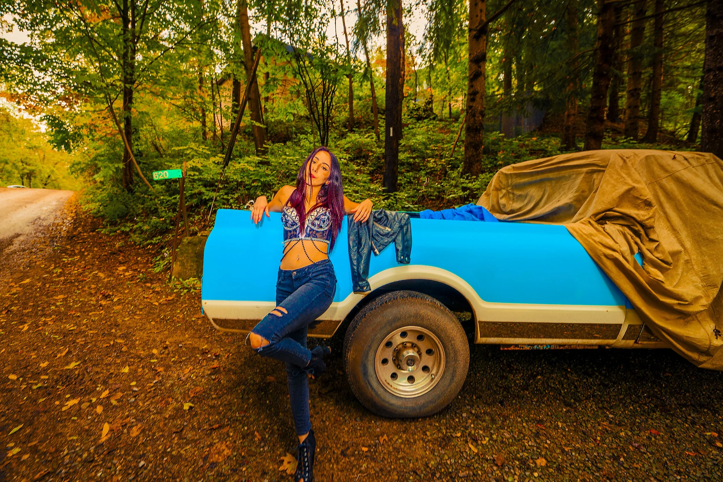 A woman with purple hair, wearing a black and purple top and ripped jeans, leaning against a blue and white trailer on a dirt road in a forest surrounded by tall trees and green foliage.