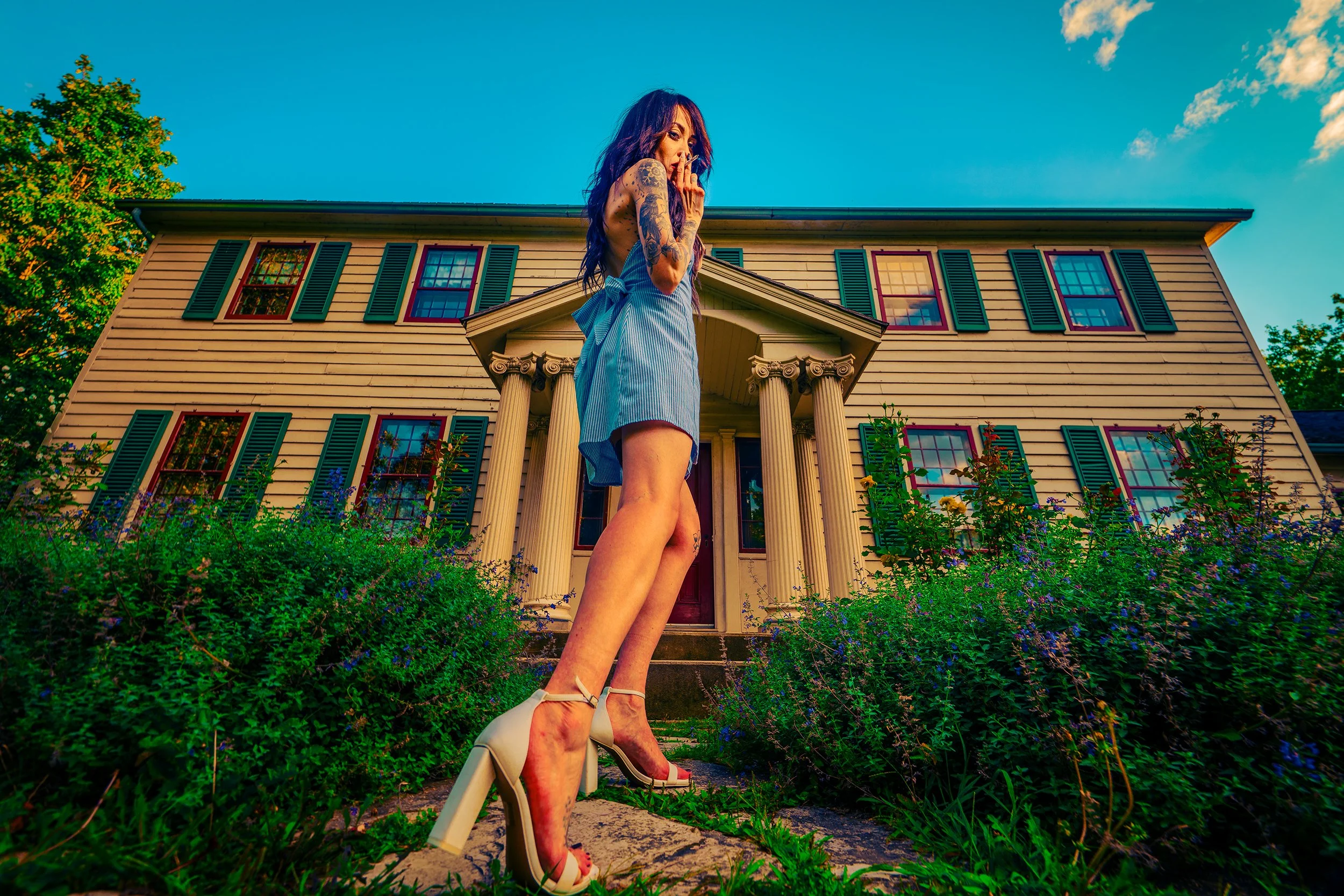 A woman with tattoos wearing a blue dress and high heels standing in front of a large house with columns and green shutters, under a blue sky with clouds.