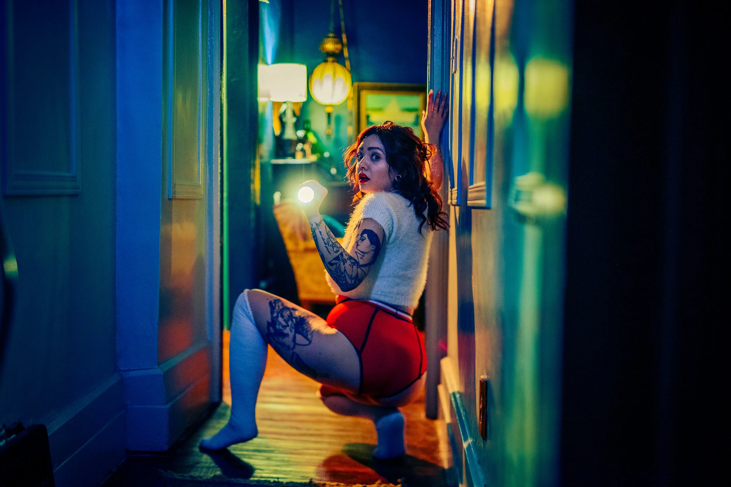 A woman with tattoos on her left arm and dark curly hair, wearing a white top and red shorts, kneeling in a narrow hallway lit by colorful lights, with her head turned to look towards the camera.