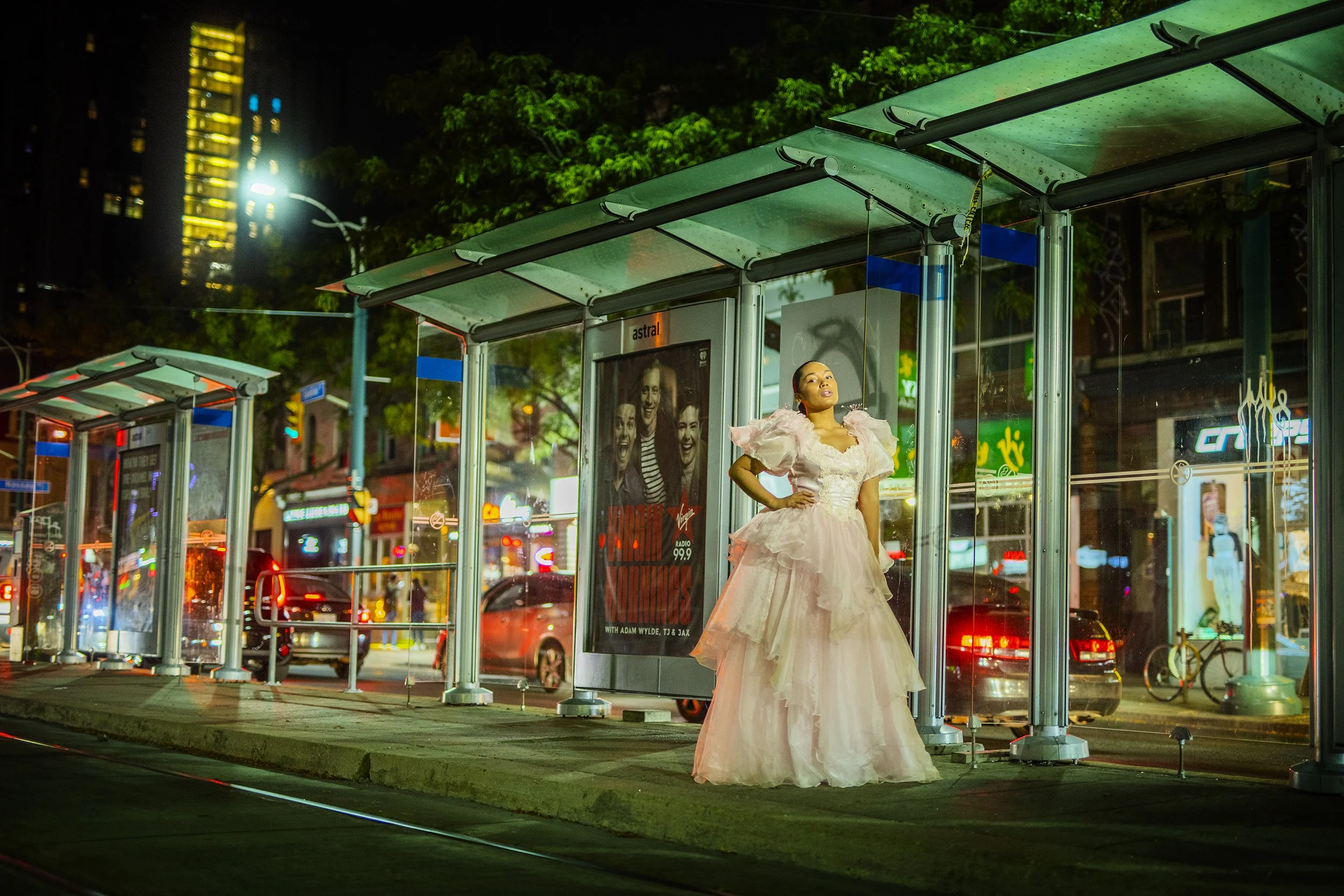 A woman in a pink, ruffled gown standing inside a glass bus shelter at night in a city setting with tall buildings and traffic lights in the background.