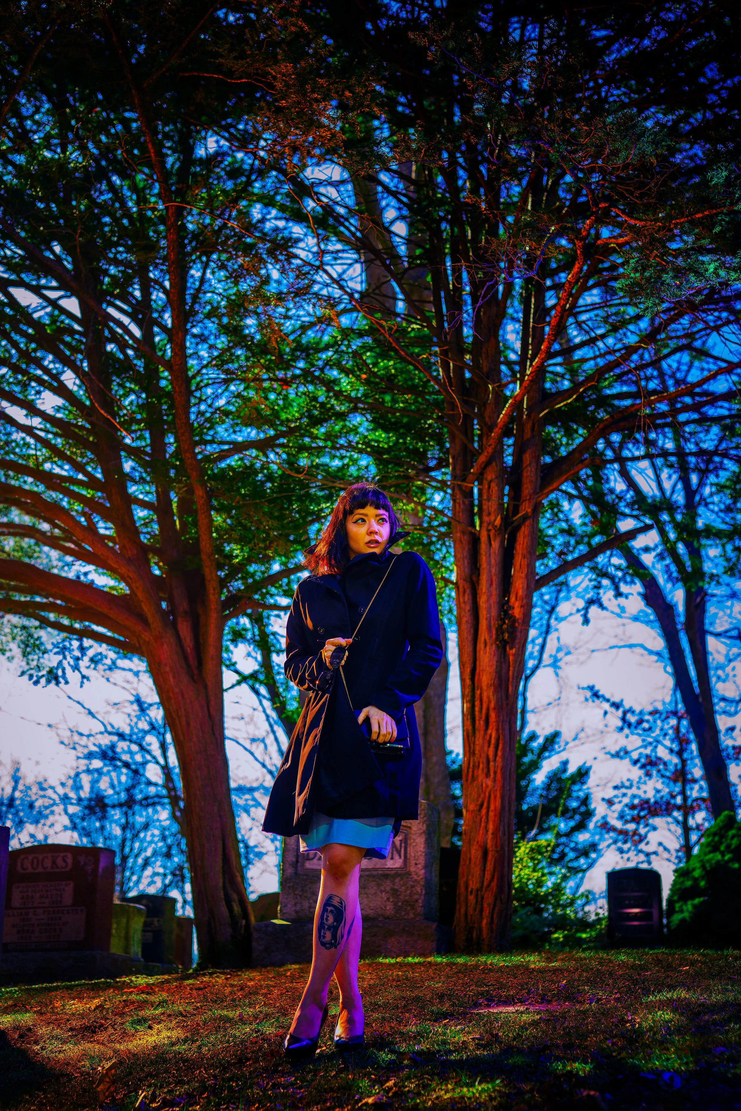 A young woman with black hair and bangs, wearing a black coat and high heels, stands on the grass in front of a large tree at dusk. She has a tattoo on her left thigh. The scene has colorful lighting, with the background showing a darkened sky and il