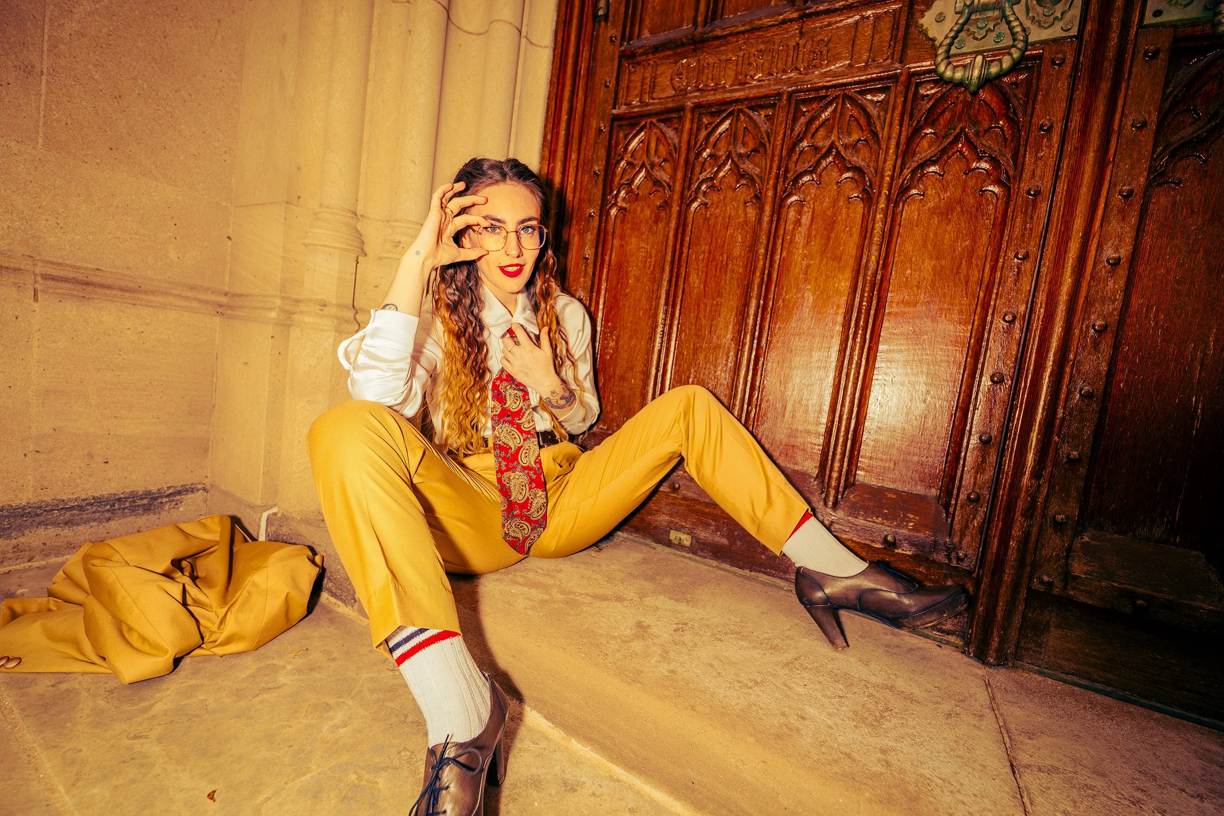 A woman sitting on the floor against a wooden door, wearing yellow pants, a white shirt, a red patterned tie, and glasses. She has long wavy hair and appears to be adjusting her glasses. There is a yellow coat or bag on the ground beside her and a br