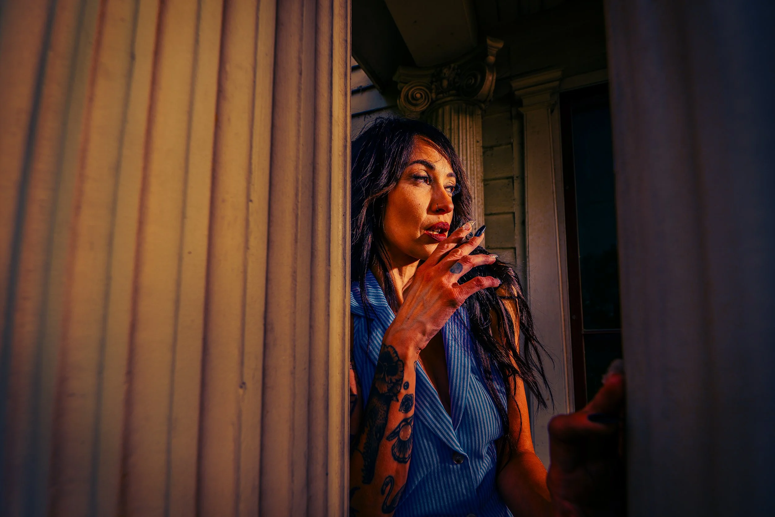 A woman with dark hair, tattoos, and makeup leaning on a window frame, smoking a cigarette inside a building during sunset.