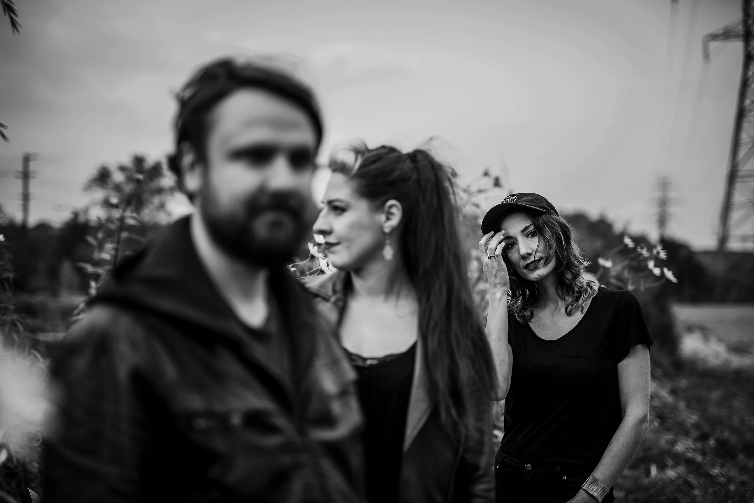 Black and white photo of three people outdoors, two women and one man, with trees and power lines in the background. The woman on the right has curly hair, wears a cap, and is looking at the camera, while the man and woman on the left are slightly ou