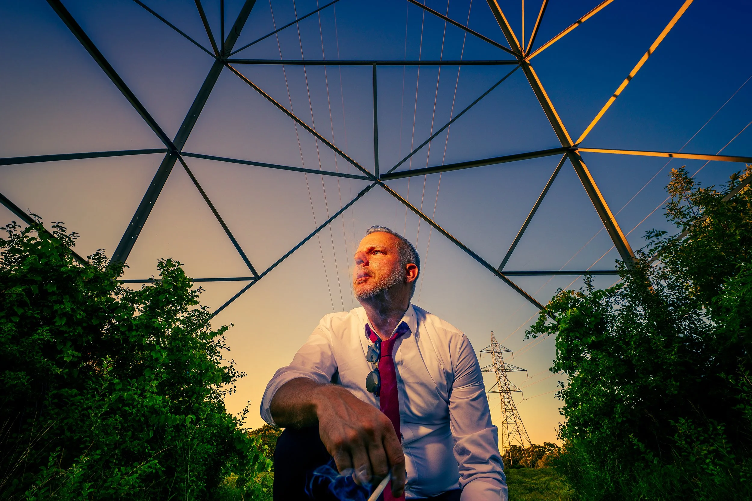 A man wearing a white shirt, red tie, and sunglasses hanging from his collar is sitting among green trees and shrubs, with a large electrical power transmission tower overhead and smaller towers in the background, during sunset.