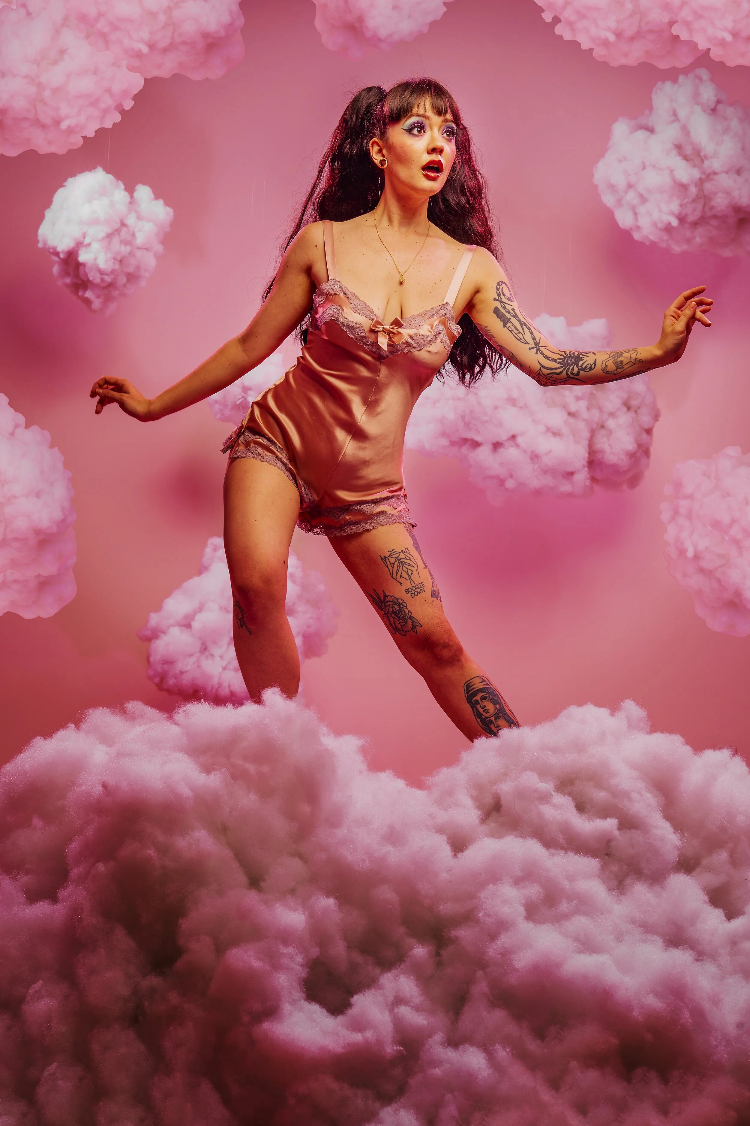 A woman wearing pink satin pajamas with lace trim standing among pink fluffy clouds and a pink background.