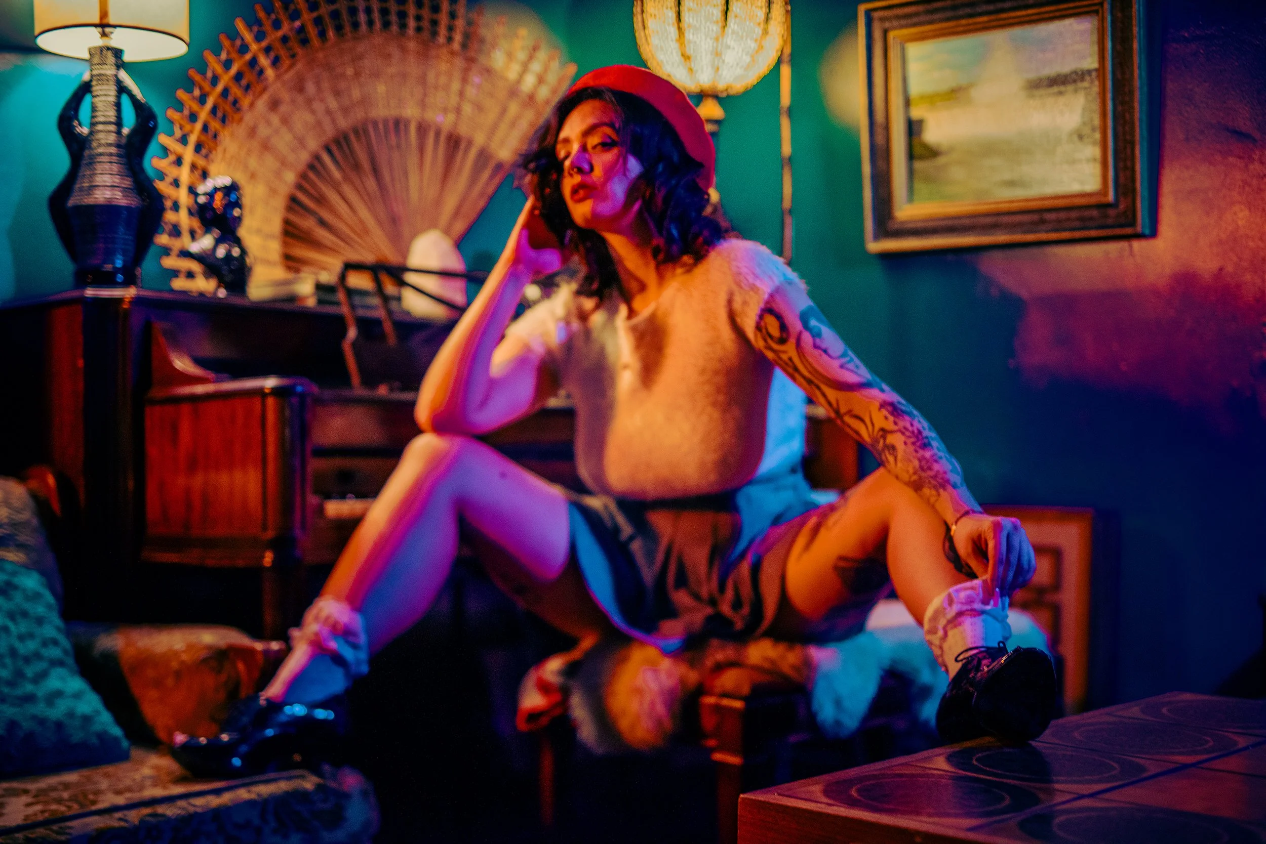 A woman with dark curly hair, wearing a red beret, sitting on the floor with one hand on her cheek. She has tattoos on her arms and is dressed in a light-colored top and patterned shorts. The room is decorated with colorful lighting, a painting on th