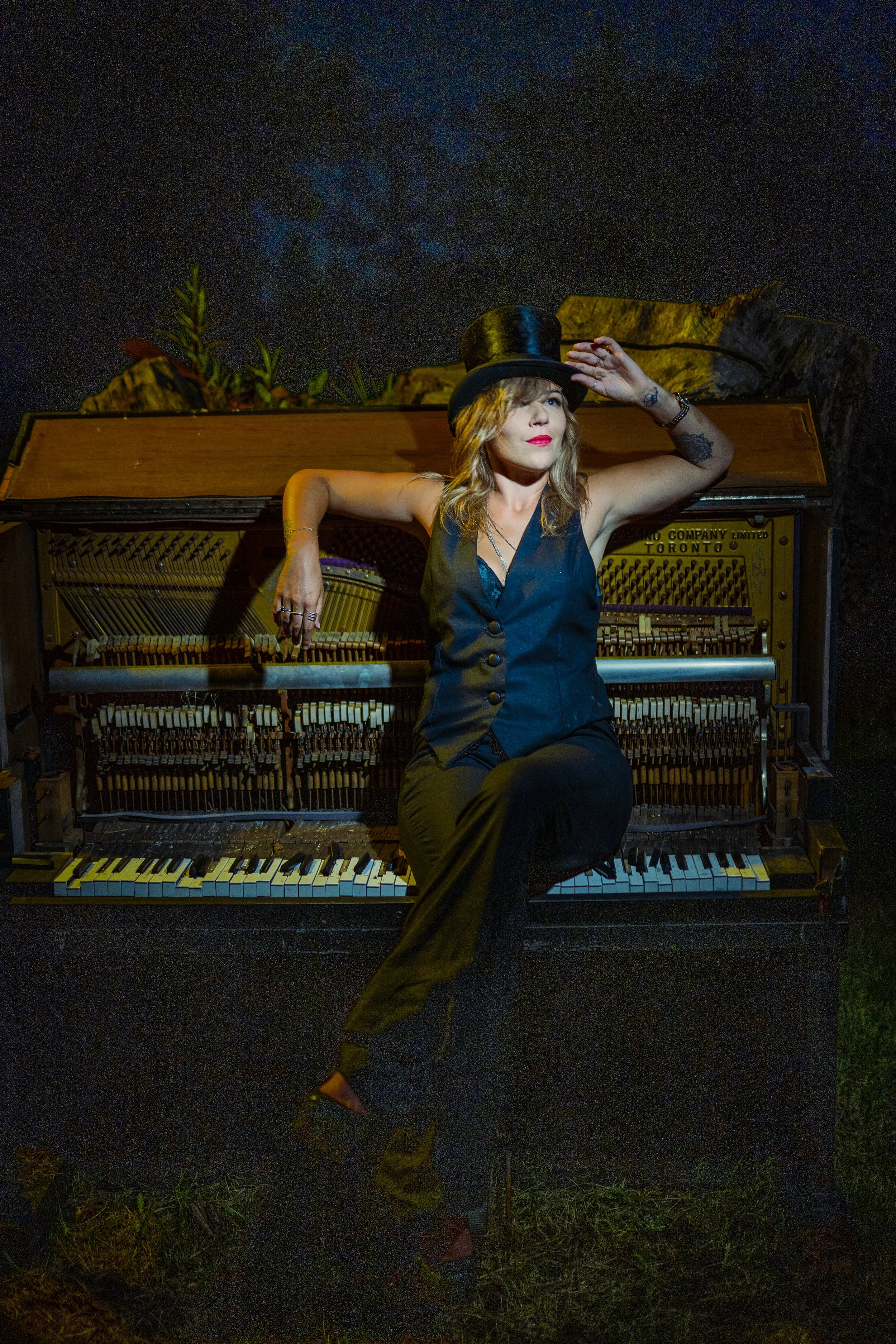 A woman with blonde hair and tattoos, dressed in black, wearing a top hat, is sitting on an upright piano outdoors at night. She is holding the brim of her hat and gazing upward. The piano's open internal strings and hammers are visible, and there ar