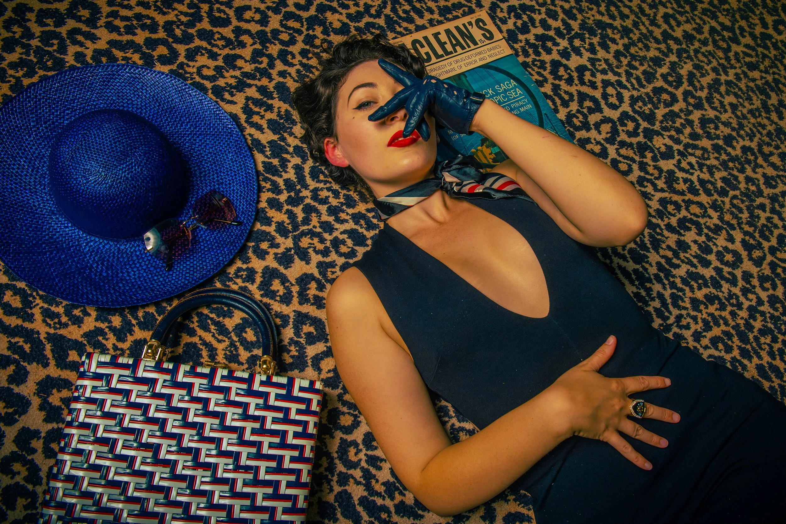 A woman lying on the floor with a leopard print carpet, wearing a black sleeveless top, red lipstick, and a patterned scarf around her neck. She is covering her nose and mouth with one hand, which is wearing a blue leather glove. Beside her, there is
