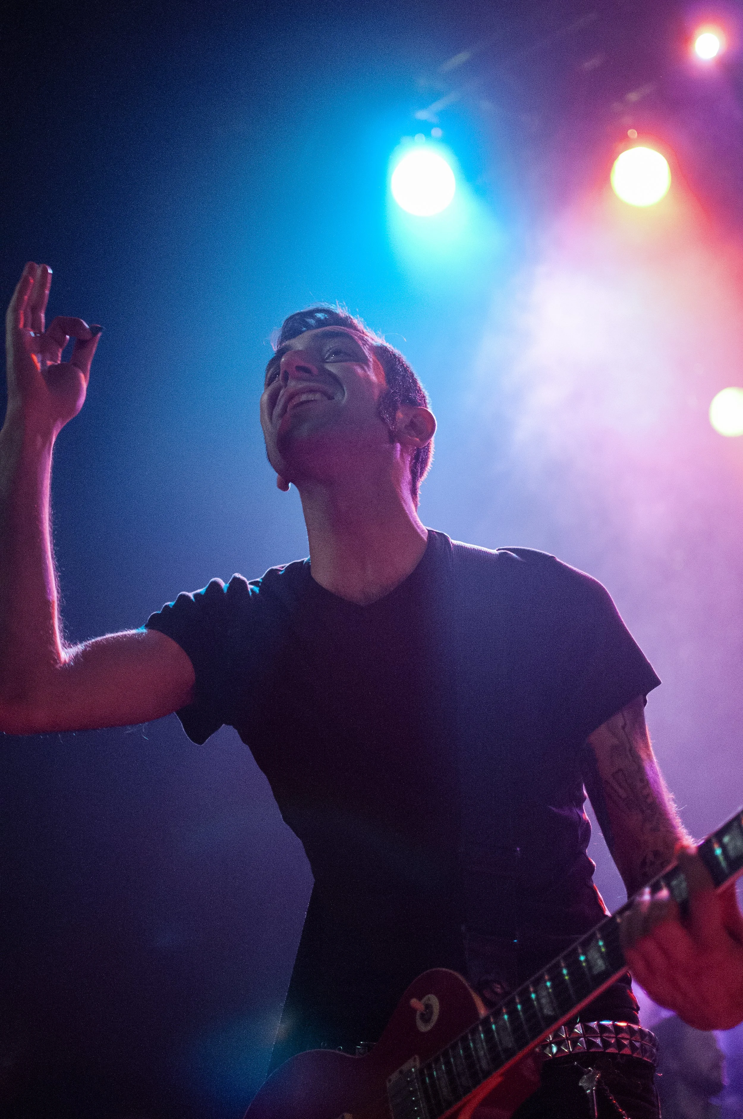 A person smiling and playing an electric guitar on stage with colorful blue, pink, and yellow stage lights overhead.