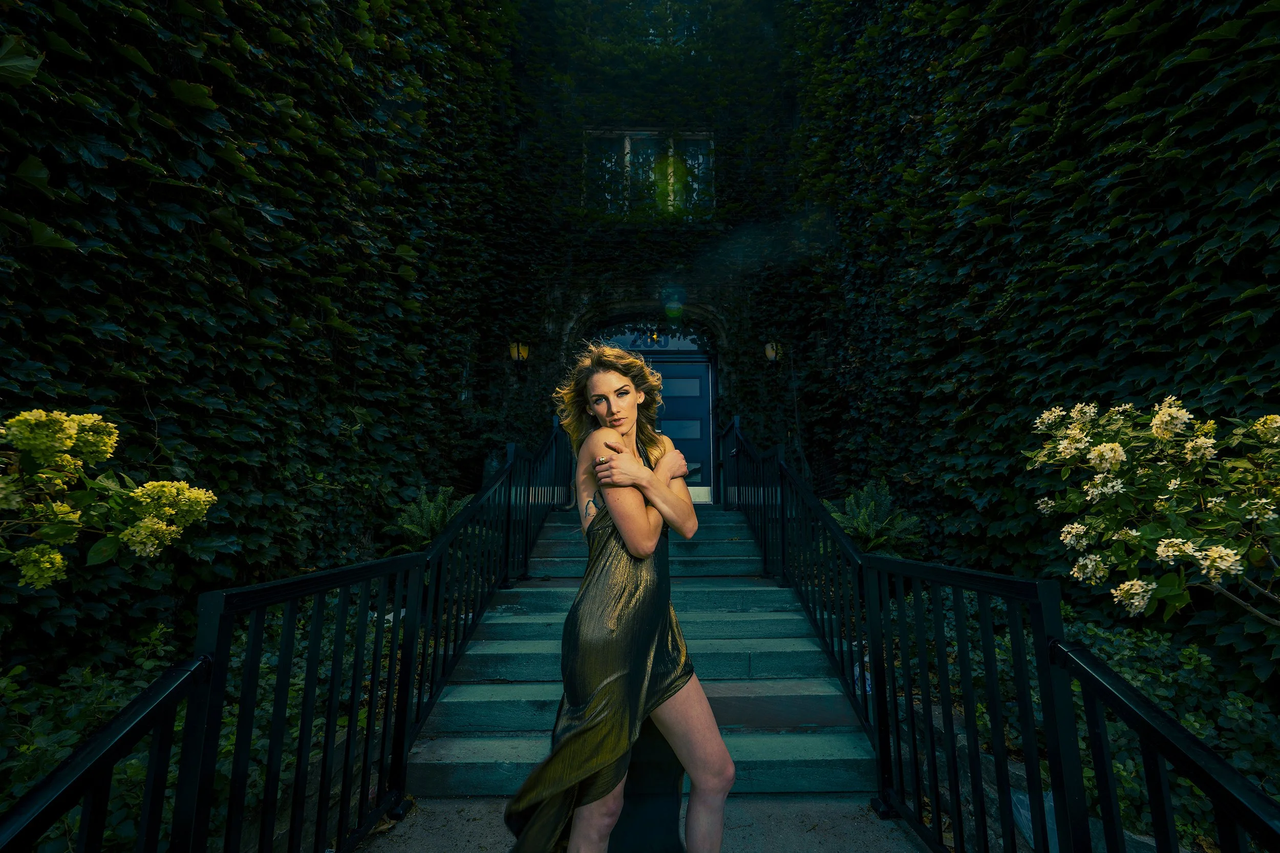 A woman in a metallic dress standing on outdoor stairs surrounded by lush greenery and flowers, with her arms crossed over her chest and looking at the camera.