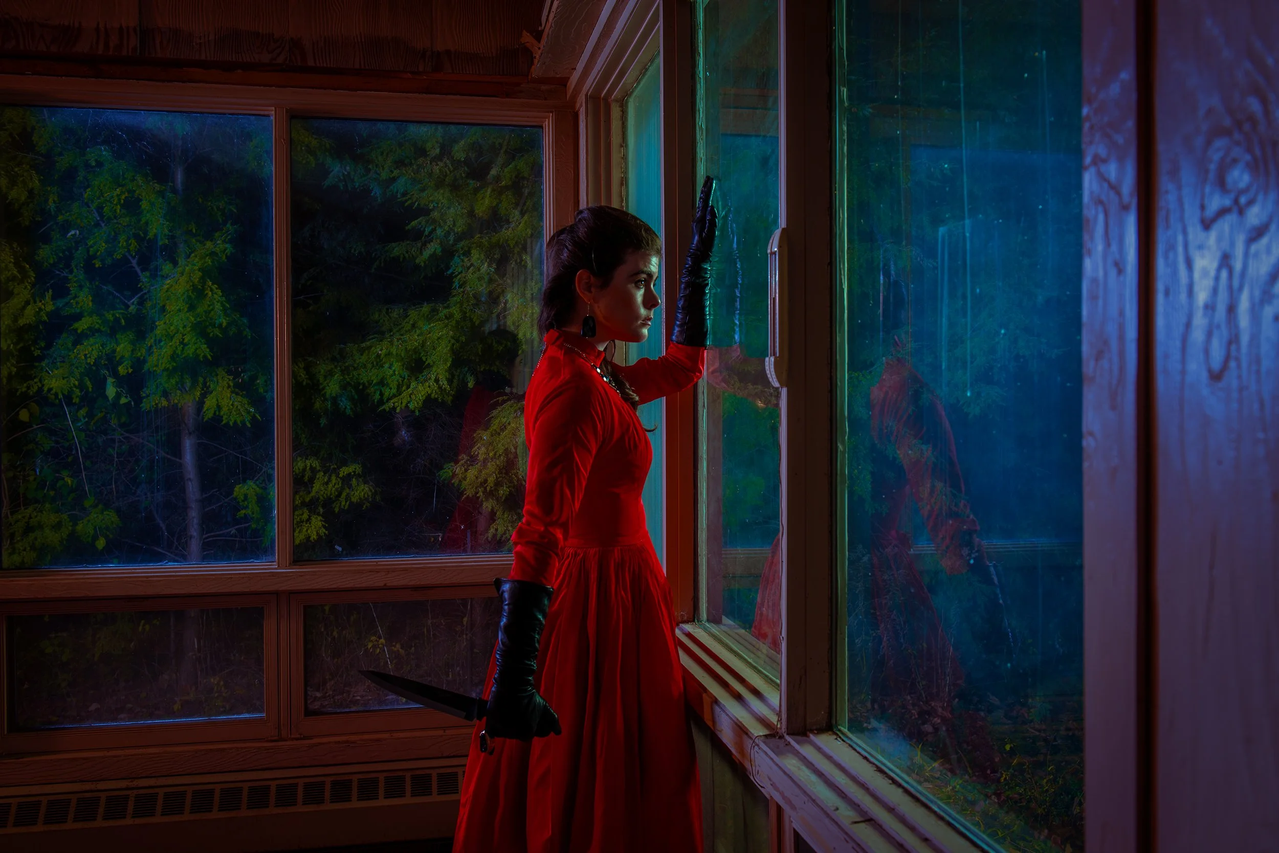 A woman in a red dress and black gloves standing inside near a large window, looking outside at a lush green forest at night.