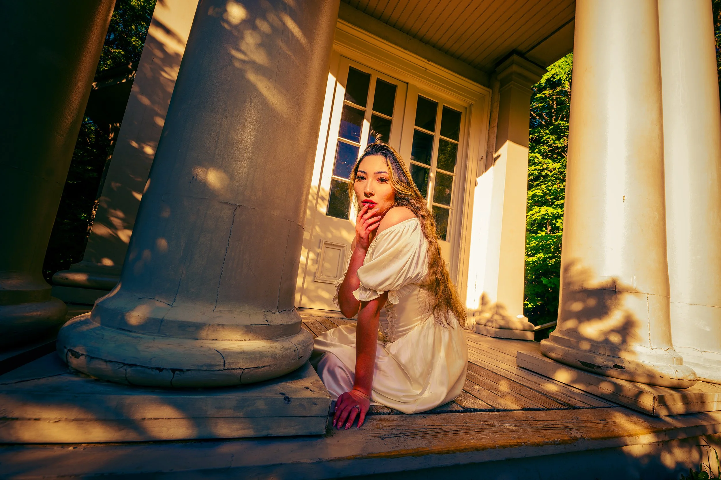 A woman in a white dress kneels on a wooden porch near large, white classical columns, with sunlight casting shadows and warm tones.