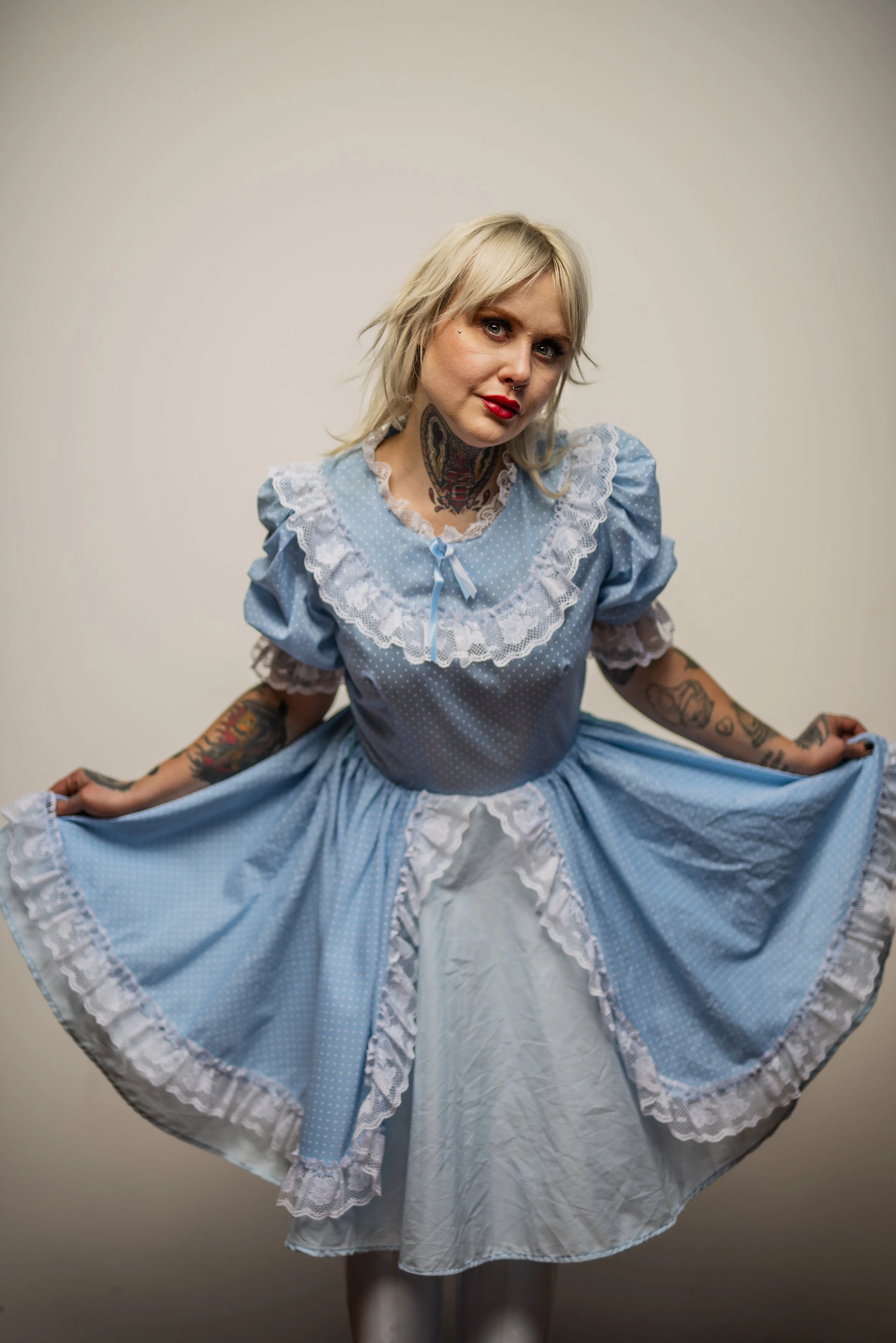 A woman with blonde hair, tattoos, and red lipstick, dressed in a blue vintage-style dress with white lace, standing in front of a plain background, holding the skirt of her dress.