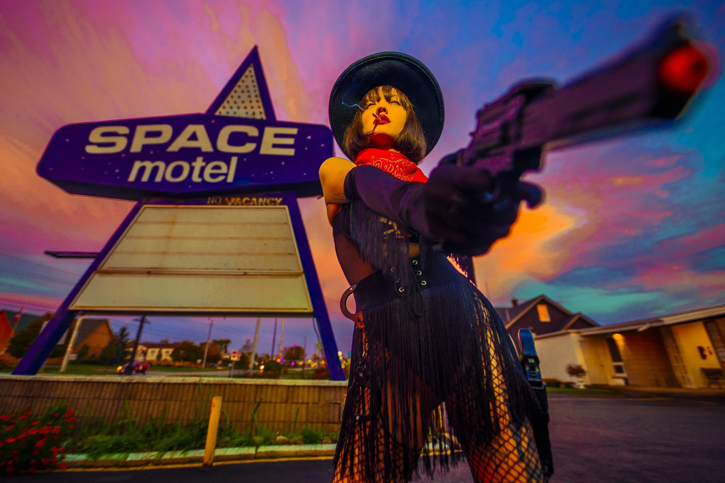 A woman dressed in a black fringed outfit with a red bandana around her neck and a black wide-brimmed hat, pointing a gun towards the camera, standing outside a retro-style space-themed motel with a colorful sunset sky in the background.