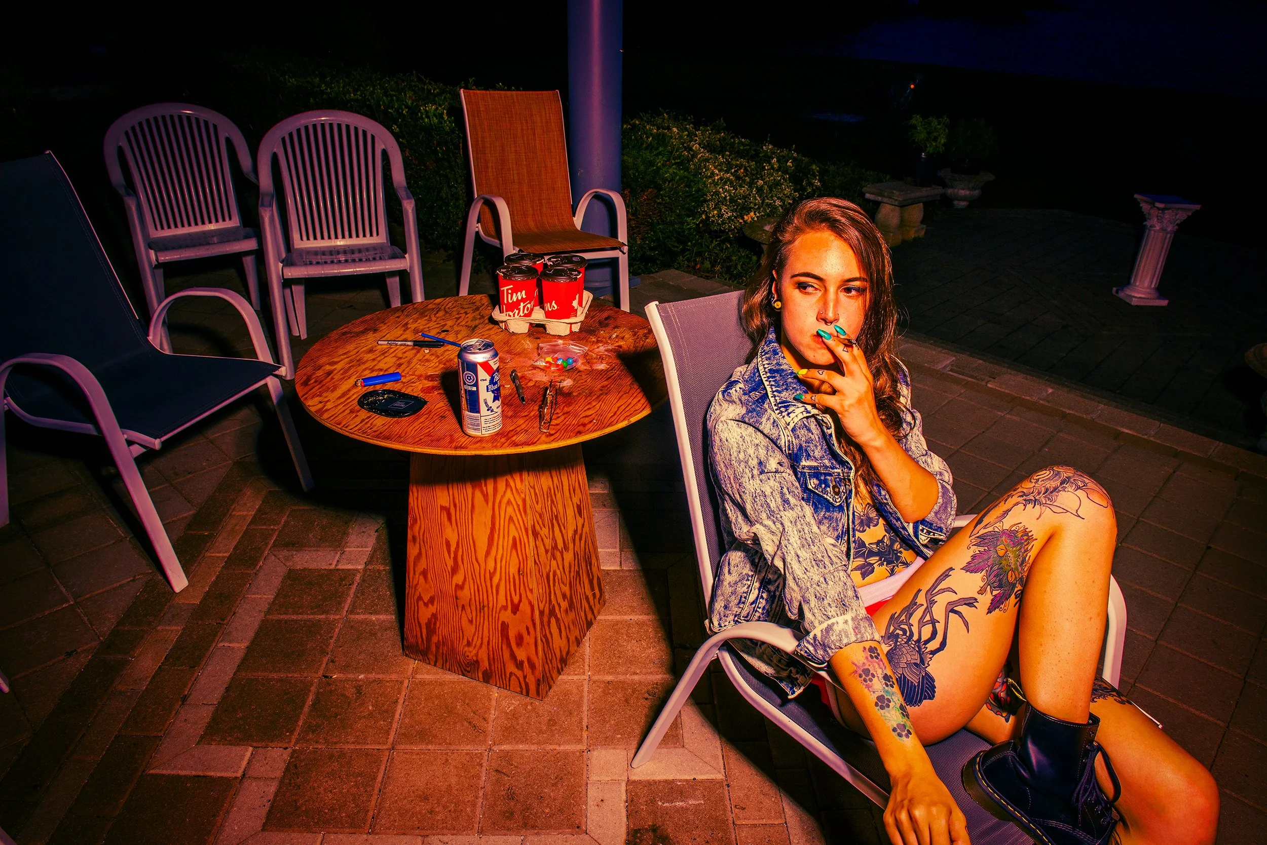 A young woman with tattoos sitting outdoors at night, with a table, chairs, snacks, and drinks behind her.