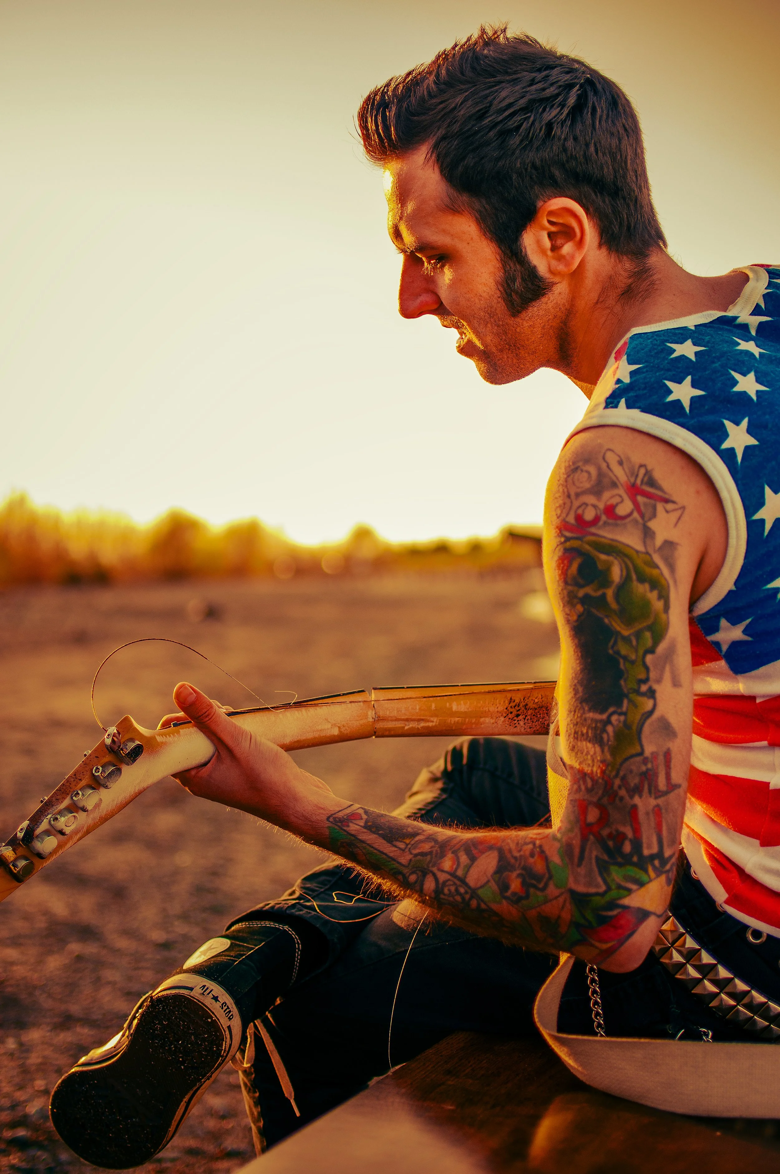 Young man with tattoos sitting outdoors during sunset, looking down at his guitar, wearing a sleeveless American flag shirt and studded belt.
