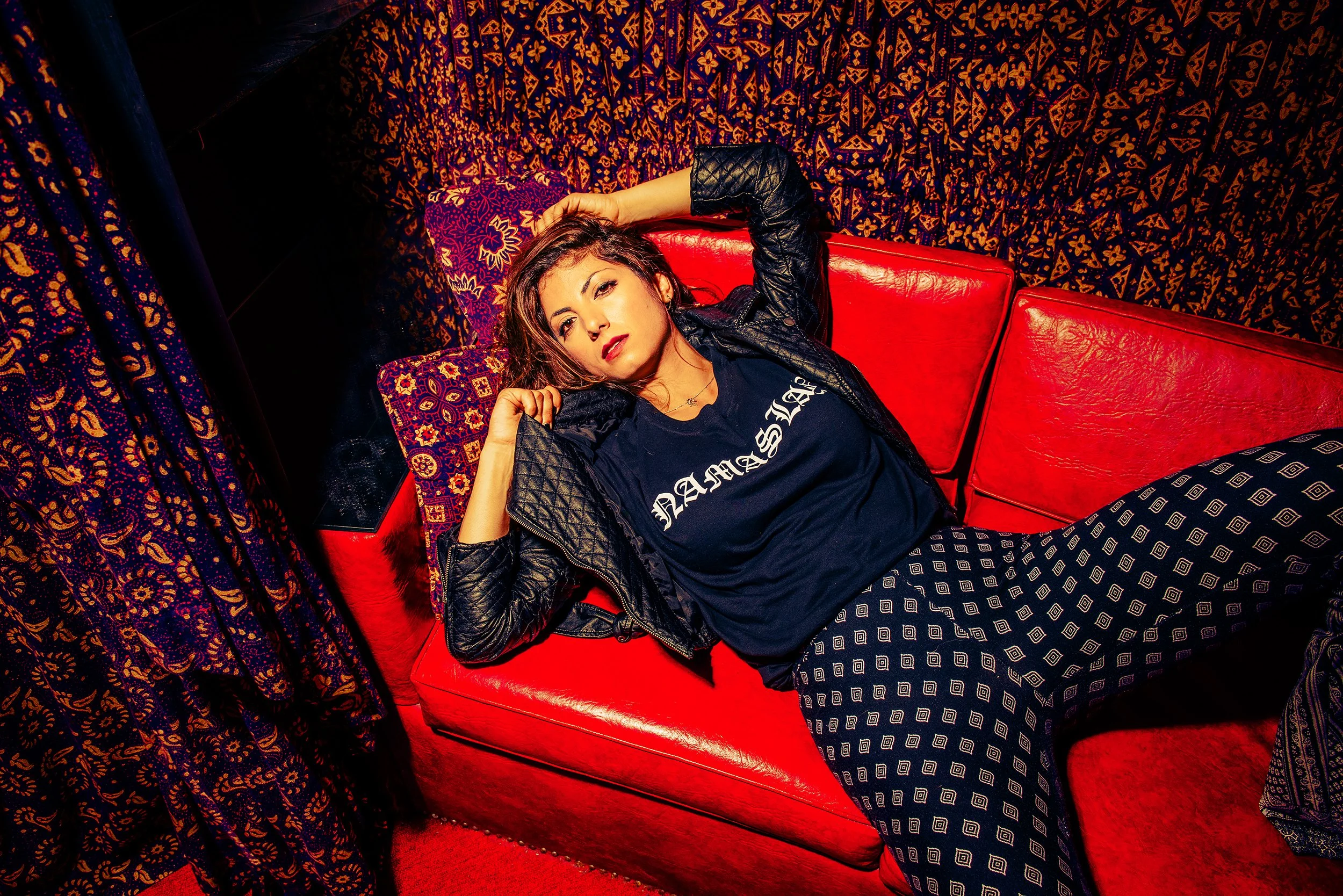 Young woman with dark hair lying on red leather couch in a room with vibrant, patterned curtains and wallpaper, wearing a black graphic T-shirt and leather jacket.