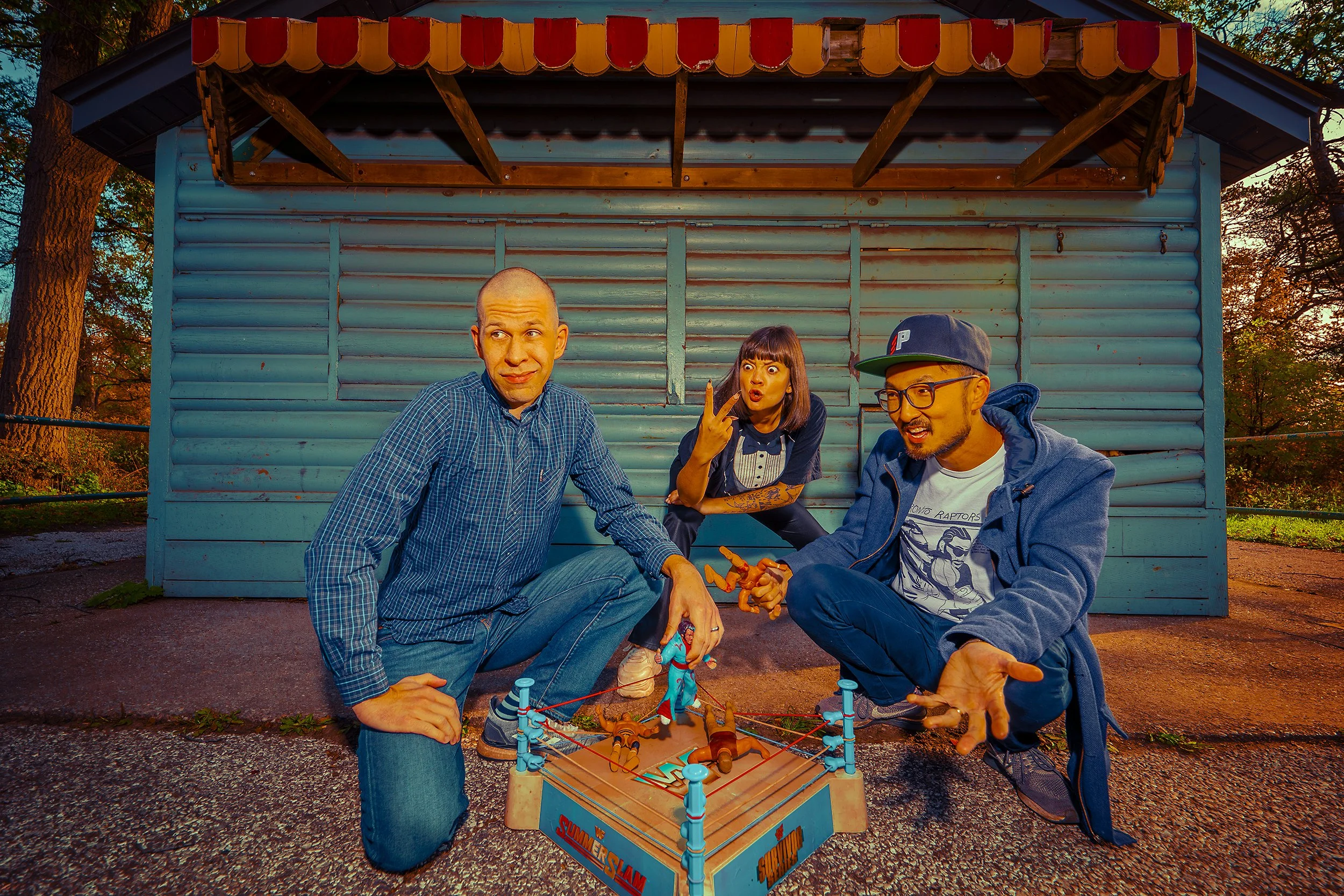 Three people playing a miniature wrestling game outside a blue wooden building during sunset