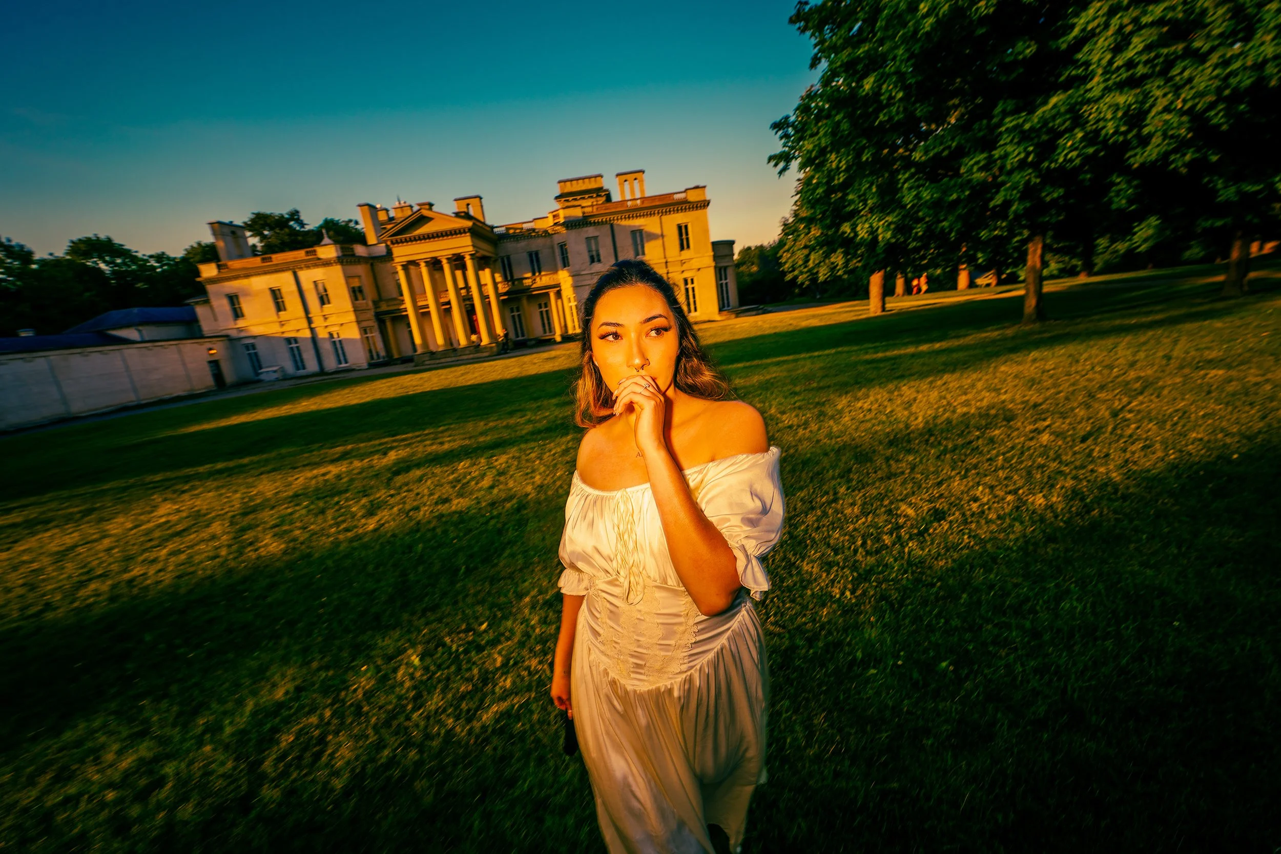 A young woman in a cream-colored dress standing on a lush green lawn in front of a large, ornate mansion during sunset.