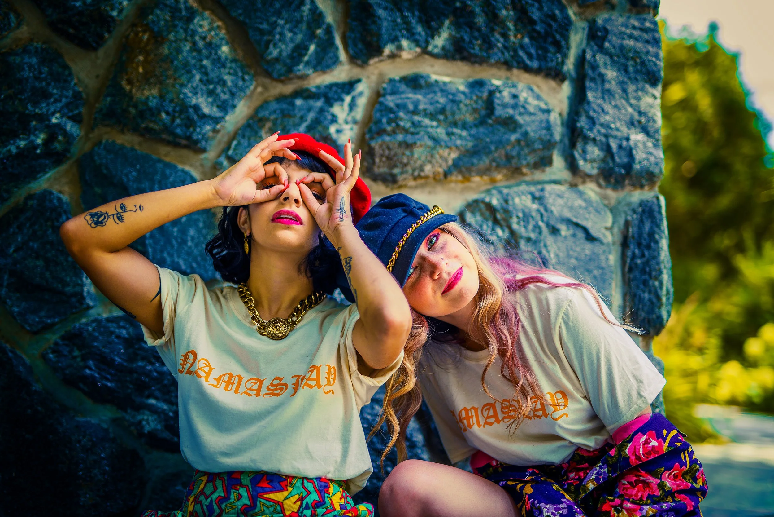 Two young women with colorful clothing and tattoos pose in front of a stone wall, with one making a hand gesture around her eye and the other resting her head on her knee.